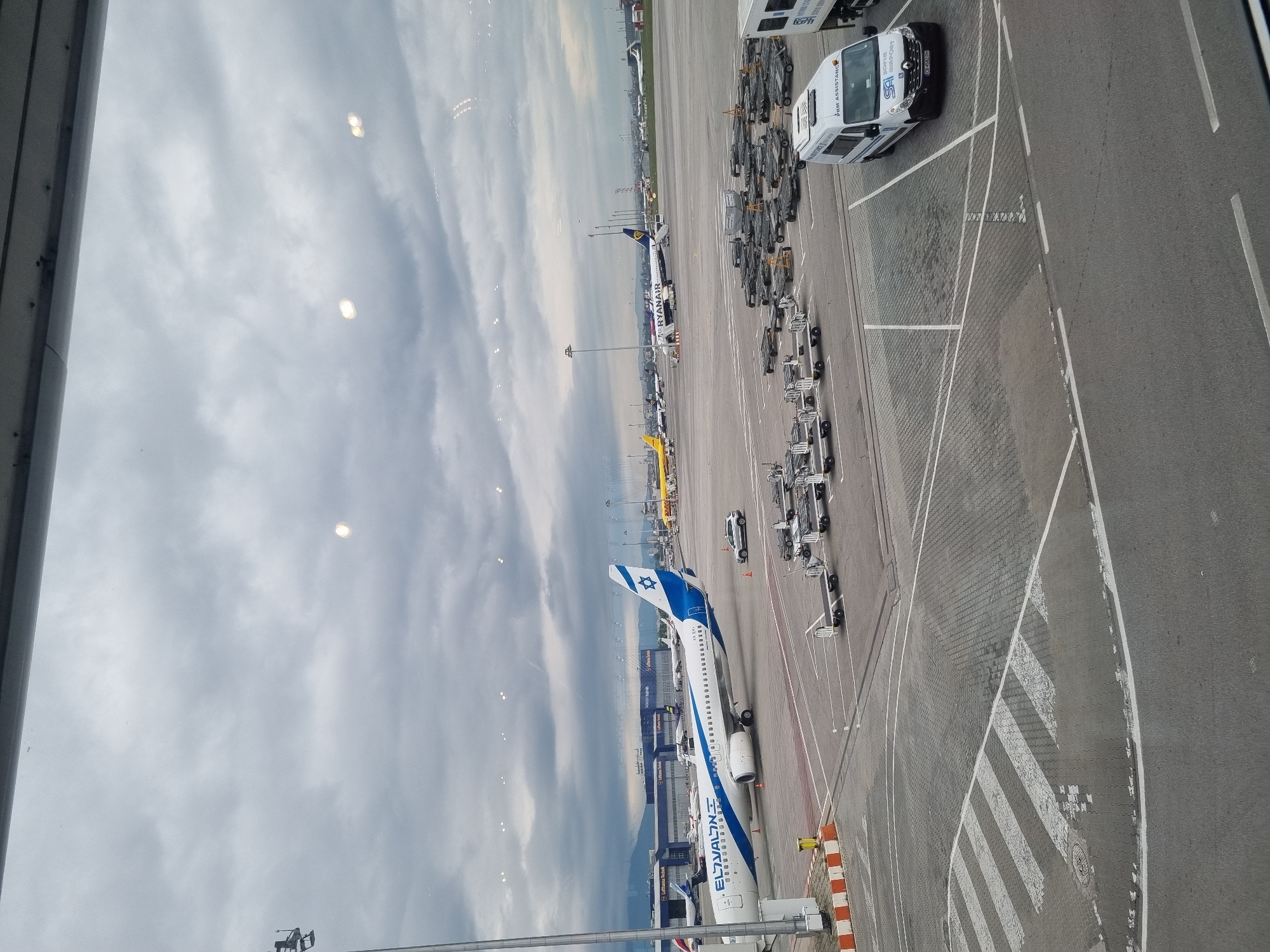 The image shows an airport tarmac, possibly at Sofia Airport in Bulgaria, as Lufthansa Technik Sofia appears in the frame. Several airplanes are visible: an El Al aircraft is prominently featured and a Ryanair plane can also be seen. Various airport vehicles and equipment, including a white van with "PRM ASSISTANCE" signage, are scattered on the tarmac. The ground surface is mainly gray concrete, with painted white lines marking designated areas. The sky overhead is cloudy.