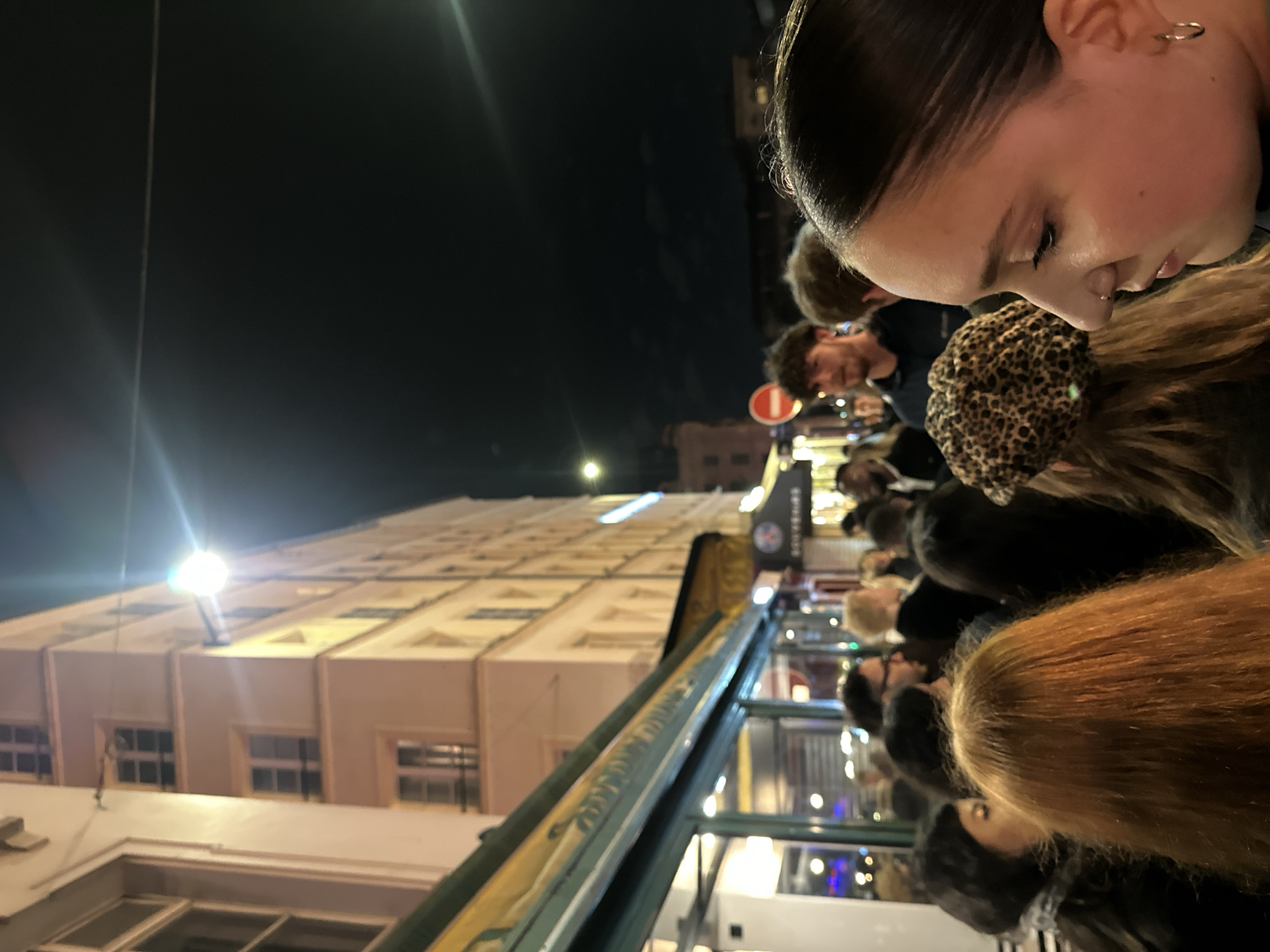 The image, taken at night, presents a view from what seems to be a moving bus or vehicle in Paris, France. On the right side of the frame, a woman with dark hair is visible, her face in profile. She has fair skin and wears a subtle piercing on her lip. The background is filled with other people, including a man with curly hair and a beard, wearing glasses. Many are standing in the bus, near windows. Outside the vehicle, an old building with light-colored architecture and rows of windows is illuminated by streetlights. The street seems to be bustling with activity. A street lamp's light is blurred at the top left of the image. A red and white street sign is visible near the top of the bus. The focus is sharp on the woman's face and the details of the architecture outside.
