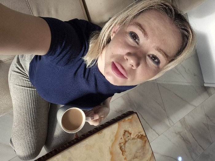 A blonde woman is captured from a high angle, wearing a dark blue top and textured grey pants. She's holding a mug of tan liquid, possibly tea or coffee, seated near a light-colored couch and marble-patterned flooring. A marble-topped table with a dark wooden trim is visible in front of her. The woman has fair skin, dark eyes, and pink lips. The lighting in the scene is bright, highlighting the different textures and patterns.