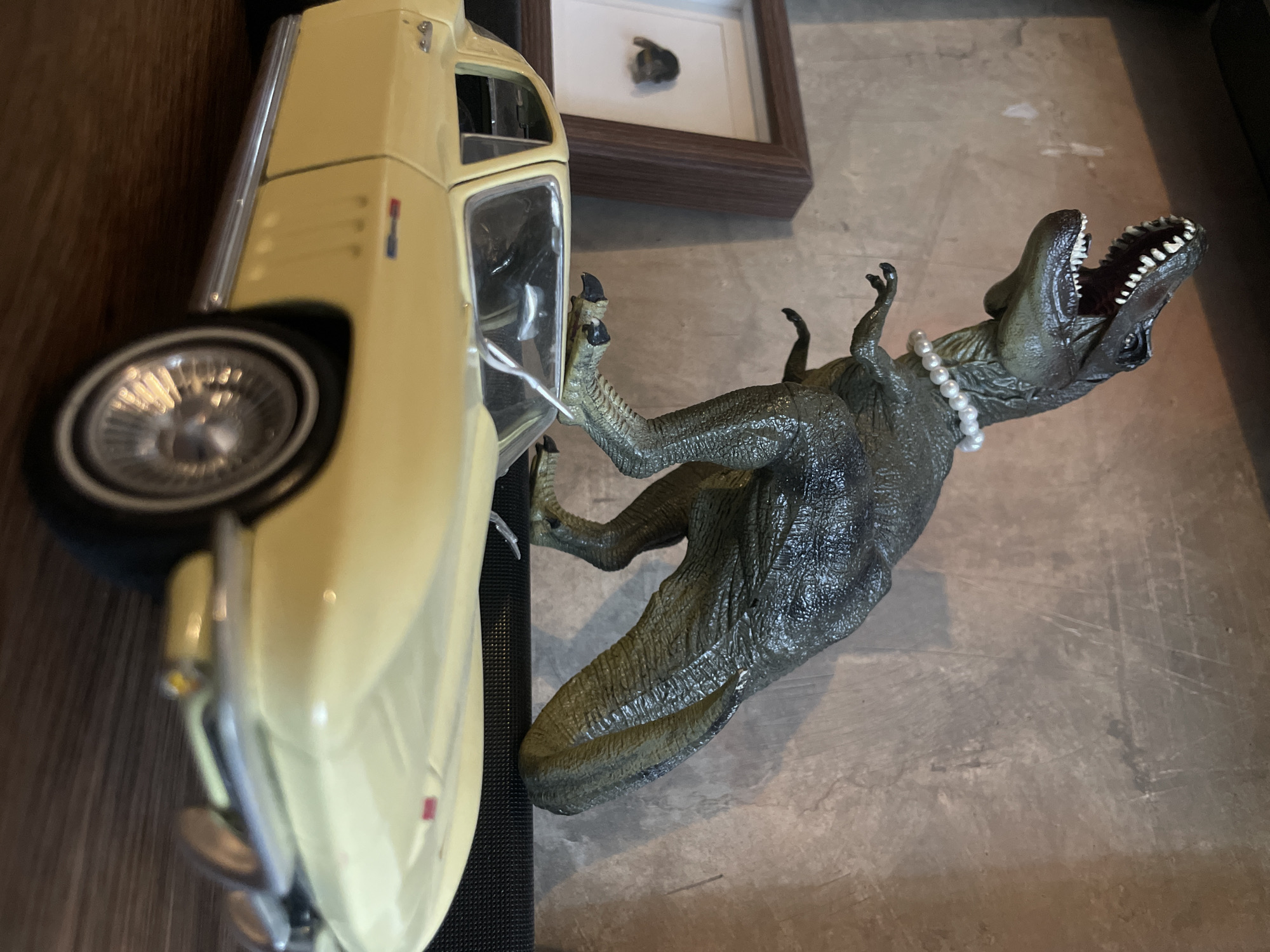 The image presents a tabletop scene featuring a yellow model car positioned vertically against a dark wooden surface, and a green dinosaur figurine with a pearl necklace oriented horizontally on a grey surface. The dinosaur appears to interact with the car, its hand making contact with the vehicle. A framed object is visible in the background. The precise location is undetermined.