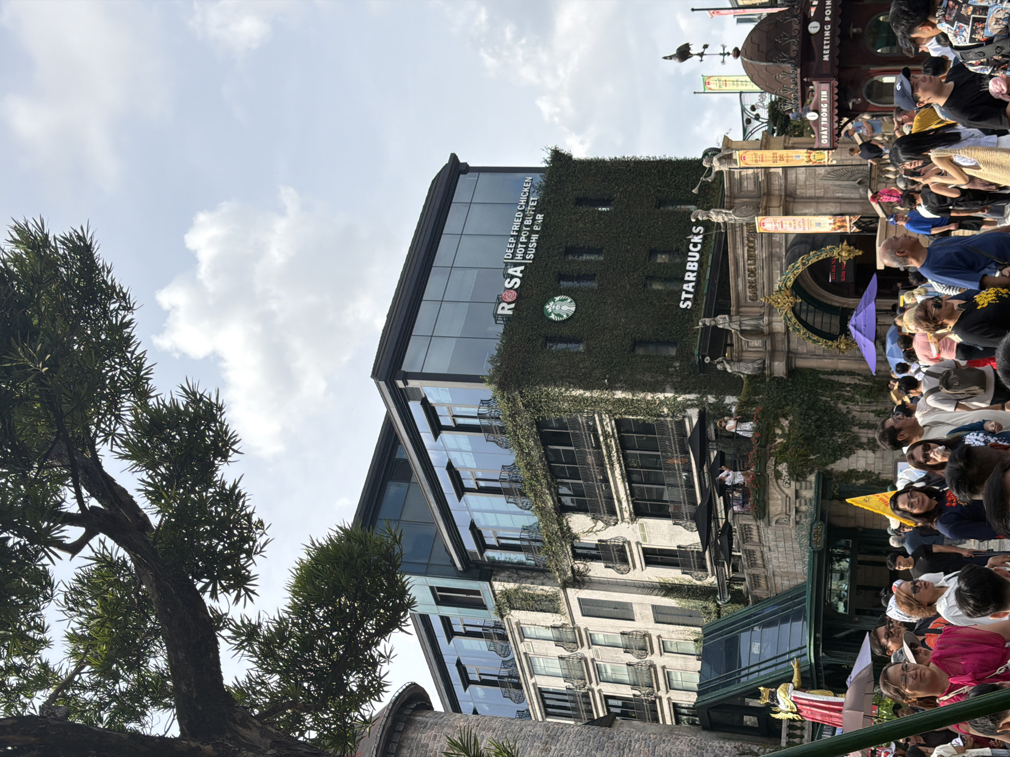 The photograph captures a bustling street scene in an urban environment, likely a tourist area. In the foreground, a crowd of diverse individuals is gathered, some looking towards the camera, others engaged in conversation or taking pictures. The architecture of the buildings suggests a blend of traditional and modern styles, with a prominent building covered in ivy, featuring large glass windows and balconies with ornate railings. A Starbucks coffee shop is visible on this building, with its iconic logo displayed. Above the Starbucks, a sign for "ROSA DEEP FRIED CHICKEN HOT POT BUFFET SUSHI BAR" is present. Further back, a fantastical-looking structure with a golden statue and a grand arched entrance adds to the unique atmosphere. The sky above is partially clouded, indicating daytime. The overall impression is one of a lively and culturally rich destination.