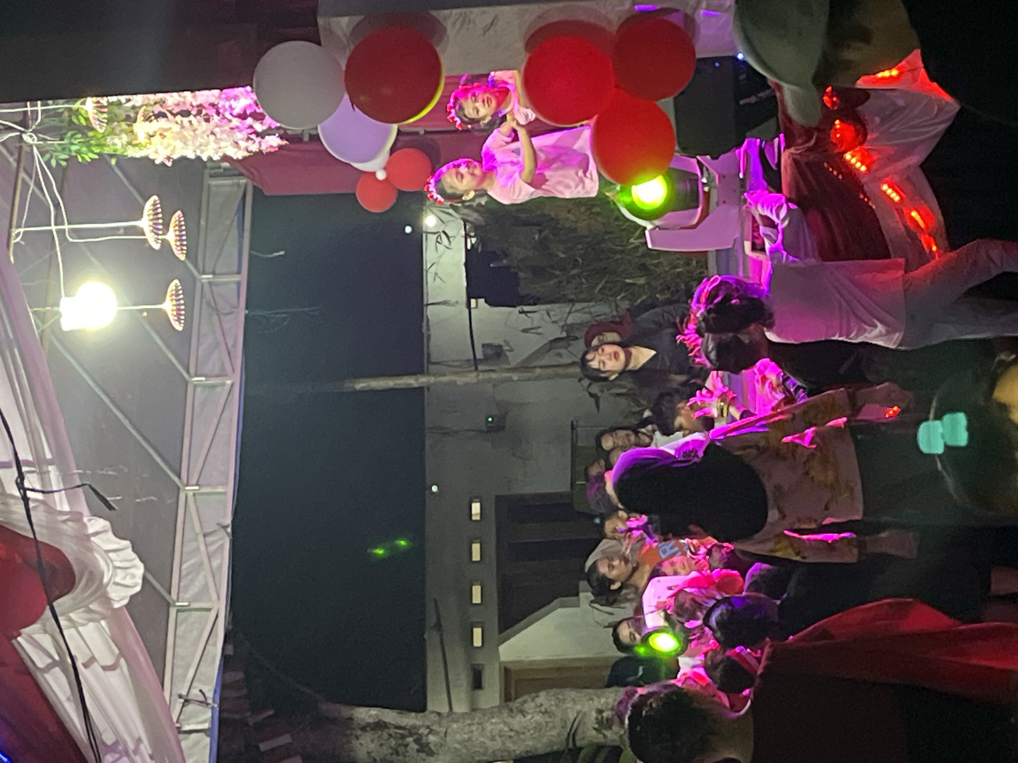 The image captures a lively outdoor event at night in Tajurhalang, Indonesia. A group of people are gathered on what appears to be a temporary stage or platform adorned with red and white decorations, possibly celebrating a national holiday given the color scheme. Two young girls, wearing pink outfits and matching red headbands, are the focal point of the stage, seemingly engaged in a performance. They are positioned near stage lights, which cast a vibrant glow on the scene.
In the foreground and surrounding the stage, a crowd of onlookers, including adults and children, are watching the event. The atmosphere is festive, indicated by the presence of balloons (red, white, and purple) and colorful string lights. The background reveals a residential area with houses, trees, and the general ambiance of a community gathering. The event appears to be illuminated by stage lights, overhead lamps, and decorative lighting, creating a somewhat dim but festive environment.