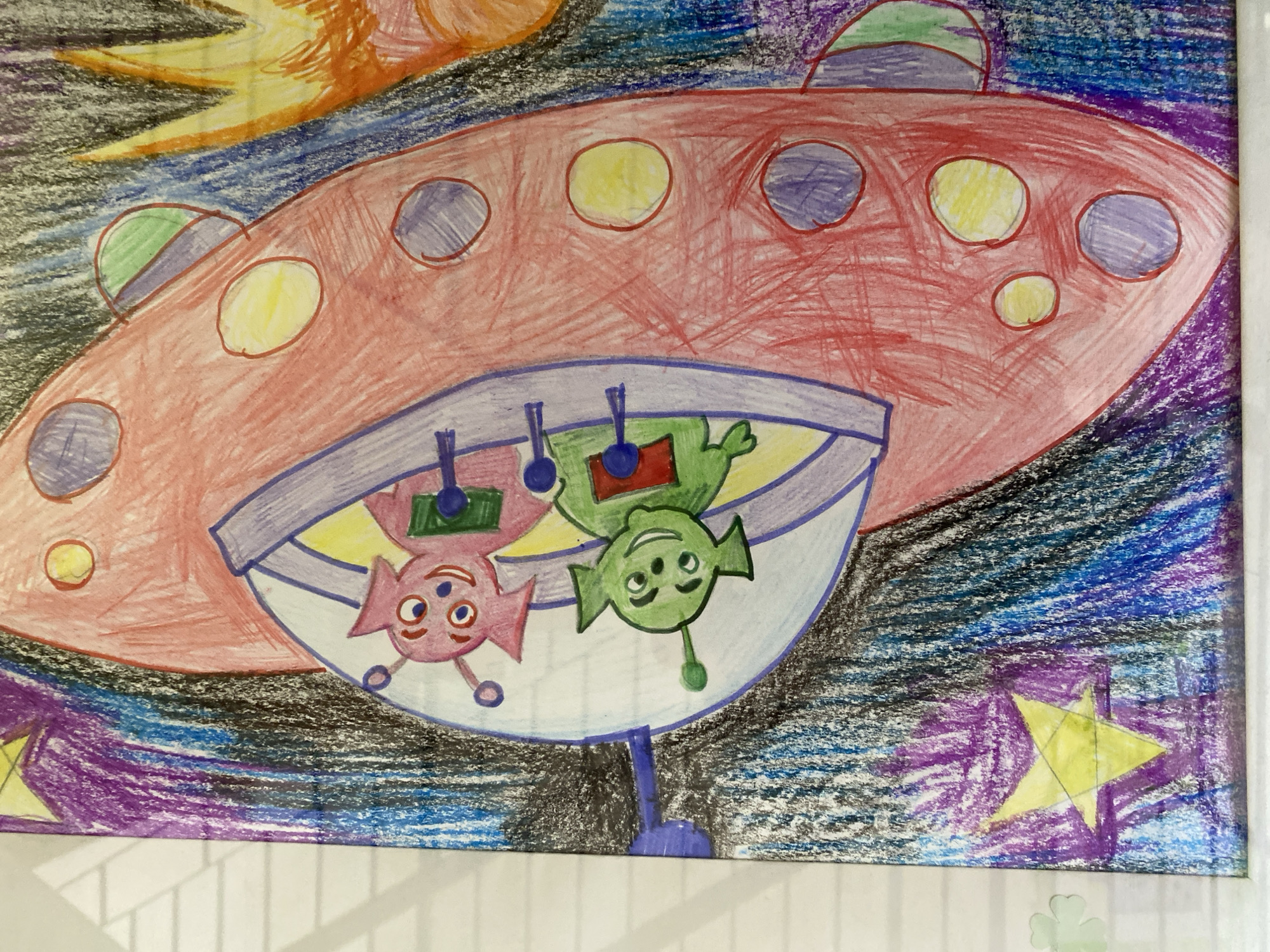 The image is a child's drawing of two aliens in a spaceship. The spaceship is red and oval-shaped with a clear dome on top, inside which the two aliens are seated. One alien is green with big eyes and a smile, while the other is pink with wide, expressive eyes and a grin. They are both wearing green outfits with blue accents. The background is a dark, starry sky with purple and blue streaks and yellow stars. There are also some circular shapes on the side of the spaceship, colored yellow and purple. A fiery orange and yellow object is visible in the bottom right corner of the drawing. The drawing appears to be done with crayons or colored pencils on paper. The location in the city of Kaliningrad, Russia is not depicted or suggested in this image.
