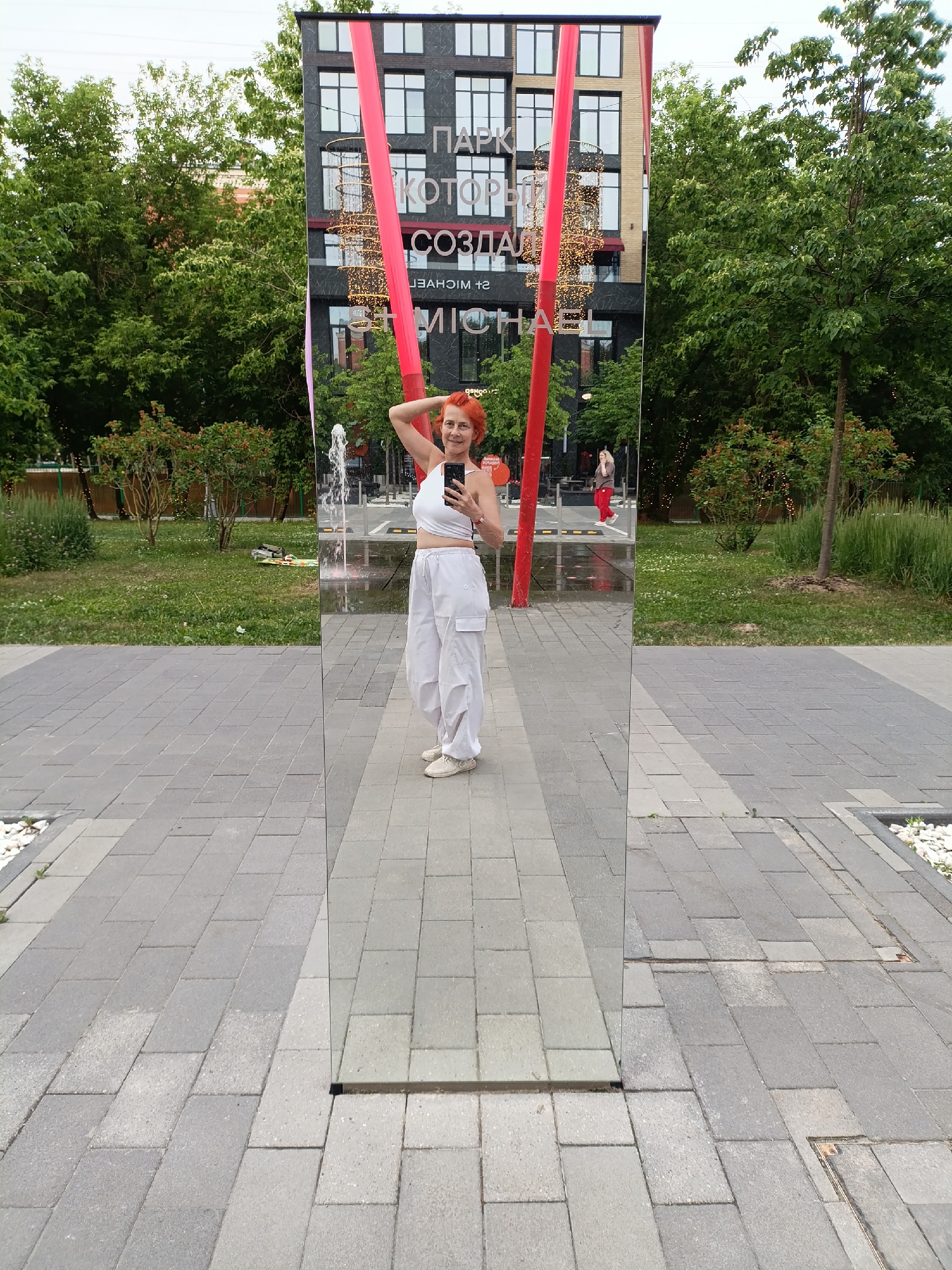 A woman with short, bright red hair stands in front of a tall, rectangular mirror, taking a selfie with her phone. She is wearing a white crop top and loose white pants. Her left arm is raised and her hand is resting on her head. The mirror reflects the surrounding environment, including a building with text on it, trees, and a fountain. The text on the building appears to be in Russian. The scene takes place on a paved walkway with a grassy area and trees to the left. The building in the reflection is dark gray with a pattern of windows and two bright red vertical beams. A string of lights is visible coiled around one of the beams. The paved area is made of gray rectangular pavers. In the background, there are people walking and a fountain spraying water. The woman and the mirror are the primary subjects. The overall impression is of a public urban space, likely a park or plaza.