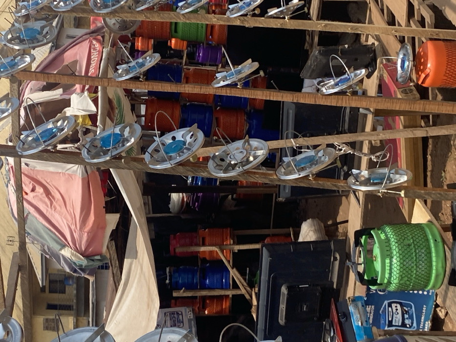 The image depicts an outdoor market stall in Afikpo, Nigeria, under bright daylight. In the foreground, a green gas cylinder with a black handle and a protective mesh is centrally positioned on a wooden surface. To its left, a blue box labeled "SYNERGY GENERATOR" supports a small, silver-colored single-burner gas stove. On the right, two vertical wooden poles are used as display stands, each exhibiting multiple small, round, silver-colored gas stove burners with blue central components attached by wires. A partial view of a person's torso and arm is visible on the far left. At the bottom right, an orange gas cylinder is topped with another small burner.
In the midground, behind the immediate display, several black flat-screen televisions are stacked. Further back, under two fabric awnings – one light-colored and another reddish-pink – numerous gas cylinders of varying colors (orange, blue, purple, and green) are vertically stacked on wooden shelves. The ground is exposed reddish-brown earth. In the distant background, a multi-story building with a light-colored facade and a brown roof is visible under a clear sky. The scene indicates a retail environment specializing in gas cooking equipment and related appliances.