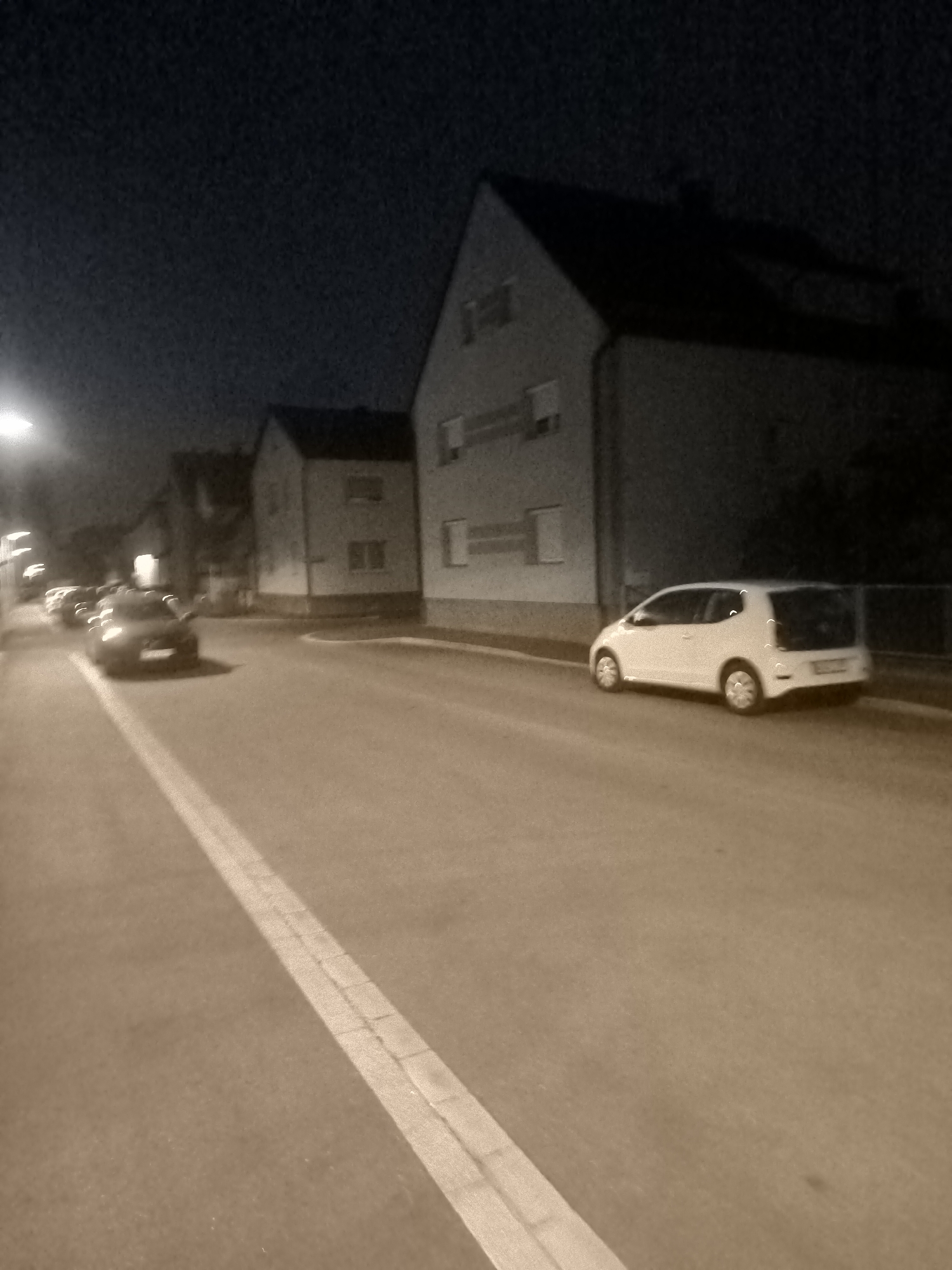 An outdoor, street-level, eye-level, long shot captures a street scene at night. The street is paved and lined with buildings on the right and a row of cars on the left. A white, compact car is parked on the right side of the street, facing away from the camera. On the left side of the street, several cars are parked in a row, with the closest one to the camera appearing to be dark in color and facing away. The buildings are multi-story structures with visible windows. The overall lighting is dim, typical of a nighttime setting, with some artificial light sources contributing to the illumination. The image has a sepia tone and a grainy texture, suggesting it was taken with a low-light camera or in low light conditions. The sky above is dark and speckled, indicative of noise from the camera sensor.