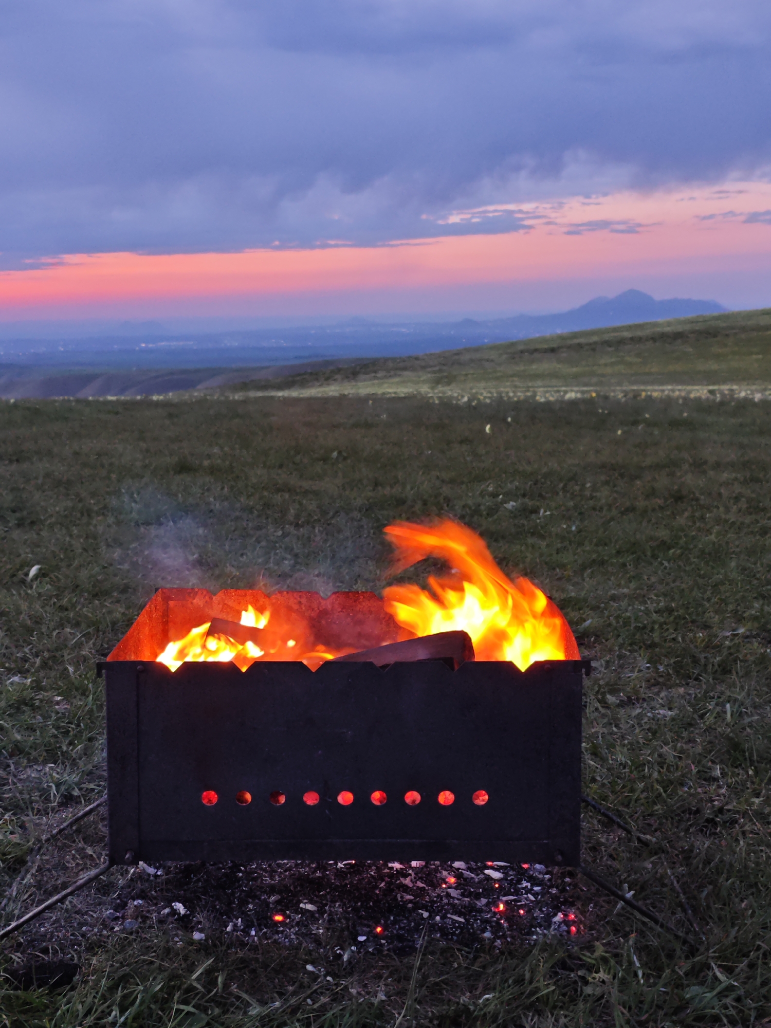 A metal grill is burning with wood in a field of grass at sunset. The grill has two rows of ventilation holes. The grass extends to the horizon, and there are mountains in the distance. The sky is mostly cloudy, with a band of pink and orange along the horizon. The location may be Lermontov, Russia.