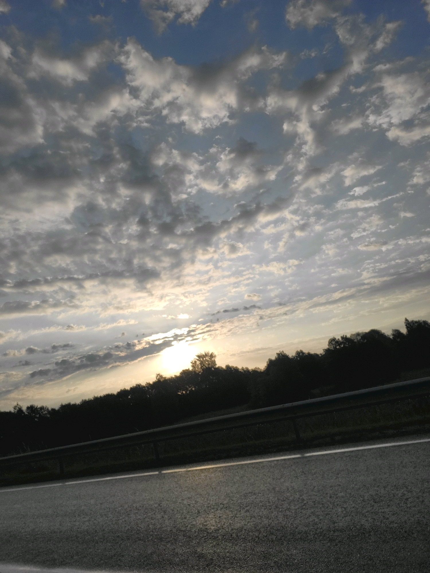 A road scene at sunset in Jelgava, Latvia, is depicted. The foreground features a dark asphalt road with a faint white line marking. A metal guardrail runs parallel to the road, separating it from a dark silhouette of trees and foliage in the midground. The sky above is filled with a dramatic display of clouds, ranging from light gray to white, with patches of blue sky visible. The setting sun is partially obscured by clouds and trees on the right side of the image, casting a warm, yellowish glow that illuminates the clouds and creates a soft light on the road surface. The overall impression is one of a tranquil, yet visually striking, natural landscape at the end of the day.