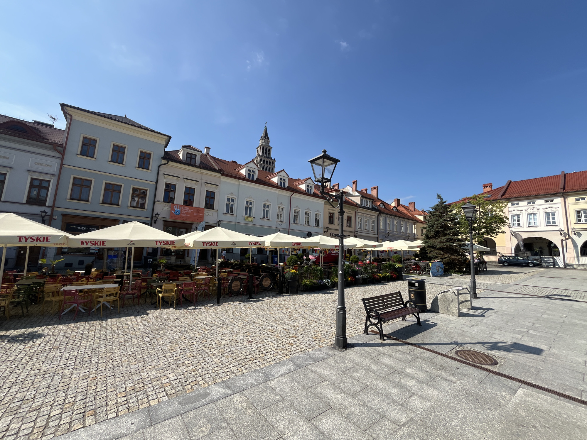 The image shows a view of a town square in front of a row of buildings, likely in Europe. The square is paved with cobblestones. In the foreground, there are outdoor seating areas with tables and chairs covered by umbrellas. The umbrellas have the TYSKIE logo on them. A black metal bench with a wooden seat is positioned on the paved area to the right of the seating. A street lamp stands near the bench. The buildings are several stories high, with pitched roofs and a variety of architectural styles. One building has a tall tower with a spire. The sky is clear and blue. There are a few people visible in the background, appearing to be patrons or staff at the outdoor seating areas. There is a parked car in the distance.