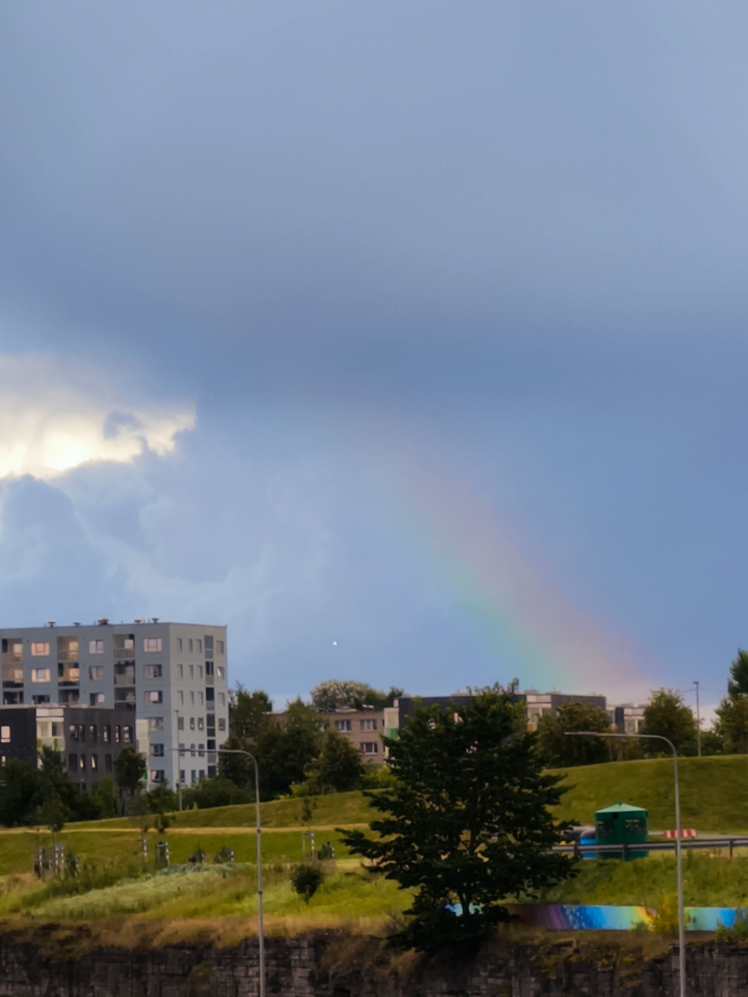 The image depicts a cloudy sky with a visible rainbow arcing across the scene. In the middle ground, several modern residential buildings with balconies and large windows are visible. A large green tree stands prominently in the foreground, casting a shadow. The landscape includes rolling green hills and a road. The scene appears to be an urban environment in Tallinn, Estonia, with the architecture suggesting a contemporary development. The lighting indicates it could be late afternoon or early evening, with the sun's rays breaking through the clouds to create the rainbow.