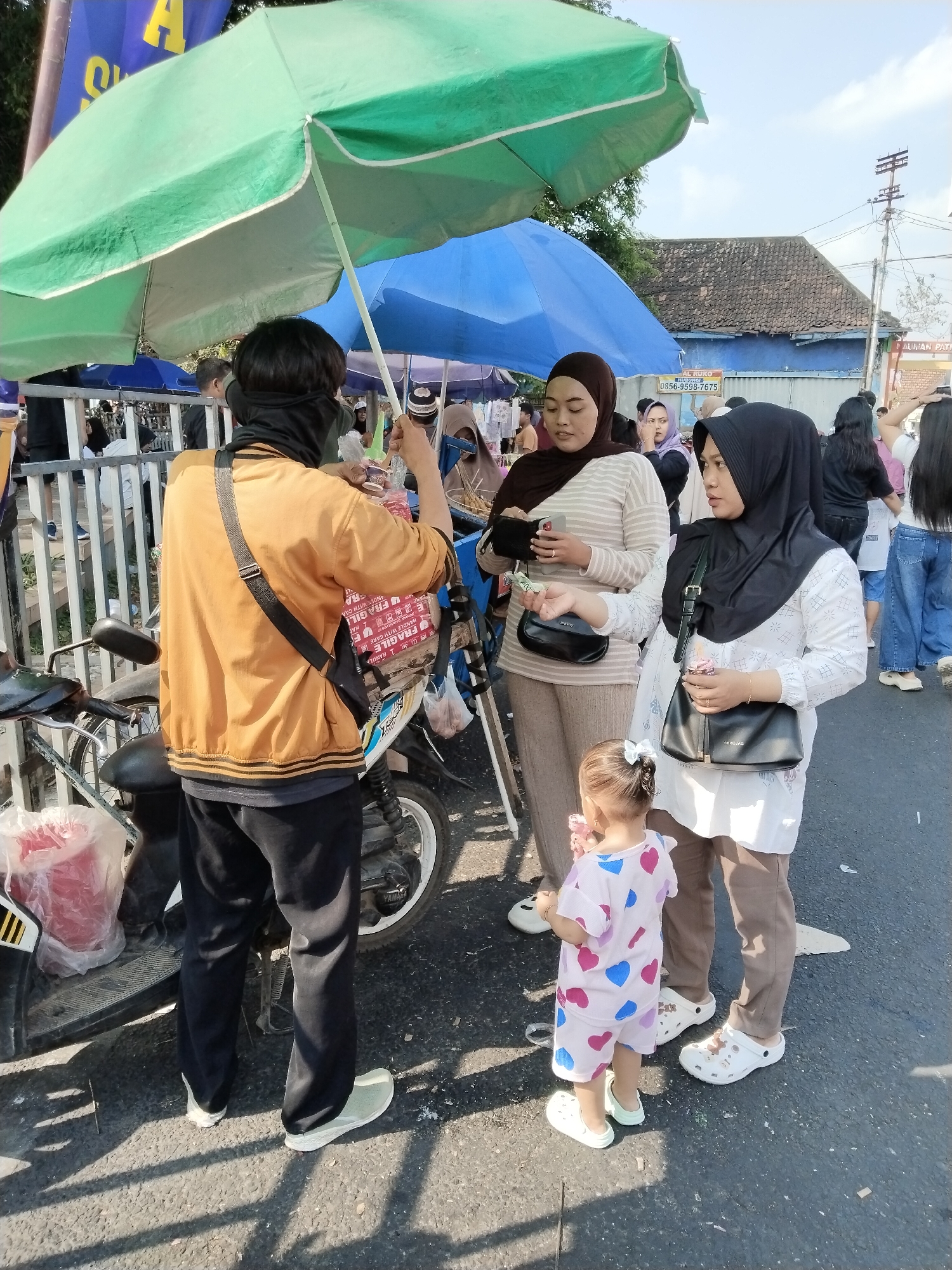 The image shows an outdoor market scene in Kecamatan Pati, Indonesia. In the foreground, a person wearing a yellow jacket and black pants stands near a motorcycle, holding a few items. They are looking down and appear to be engaged in a transaction or handling goods. To their right, two women are standing. The woman on the left, wearing a brown hijab and striped shirt, is holding a smartphone. The woman on the right, wearing a black hijab and white patterned shirt, is holding a small pink item and looking towards the person in the yellow jacket. A young child, wearing a pink and blue heart-patterned outfit, stands between the two women, also holding a pink item. In the background, there are more people, stalls, and market umbrellas, suggesting a busy marketplace. The environment appears to be a street with some buildings and utility poles visible.