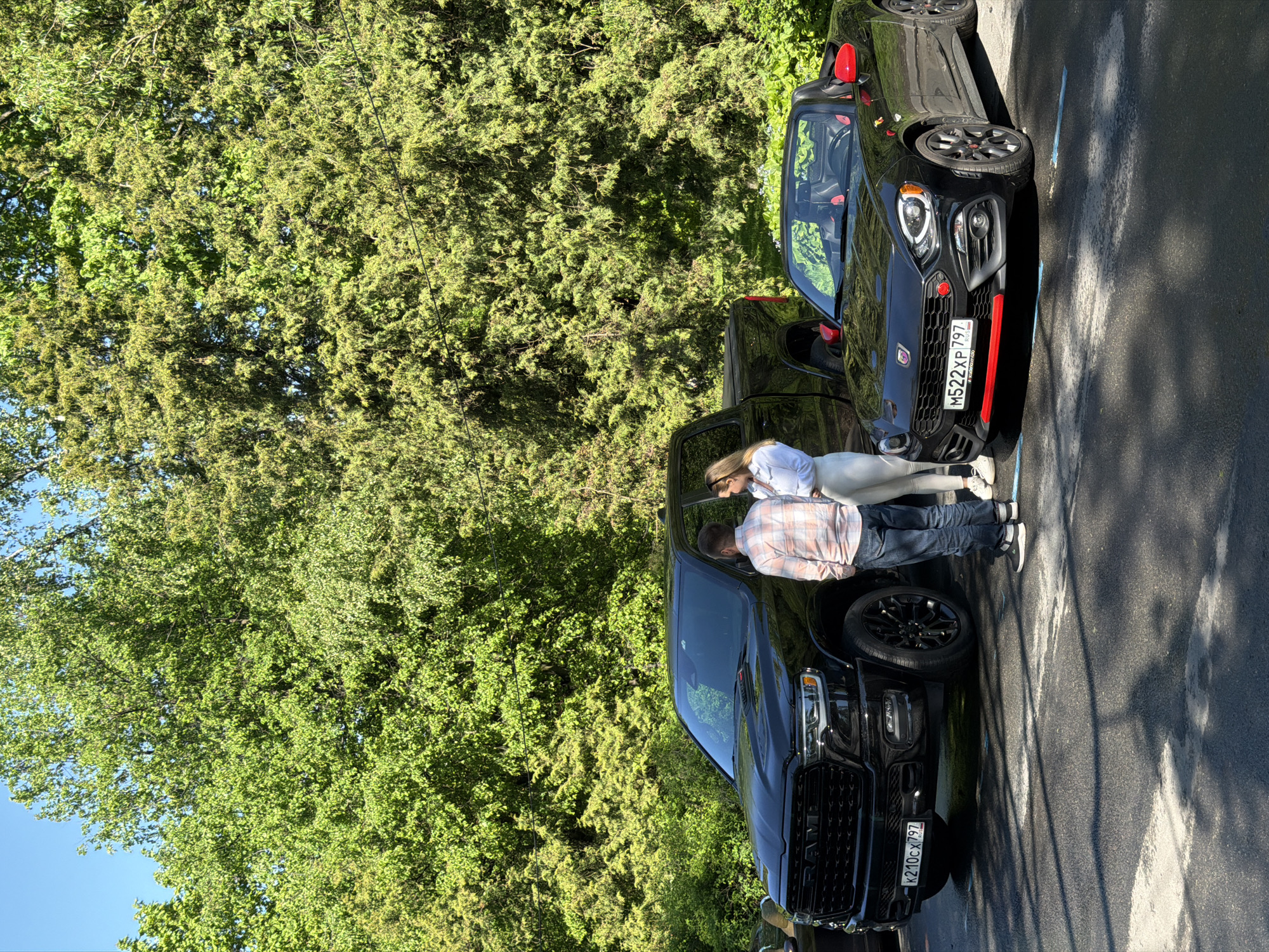 A medium-shot photo shows a man and a woman next to a dark car on a paved surface outdoors. A smaller black convertible is parked behind the truck. The license plates on both vehicles are Russian. The plates on the truck read “K210CX 797”. The plate on the sports car reads “M522XP 797.” The man wears a light pink and white plaid shirt and blue jeans, while the woman wears white. The background consists of dense green trees and a clear blue sky. This photo was most likely taken in the city of Saint Petersburg, Russia.