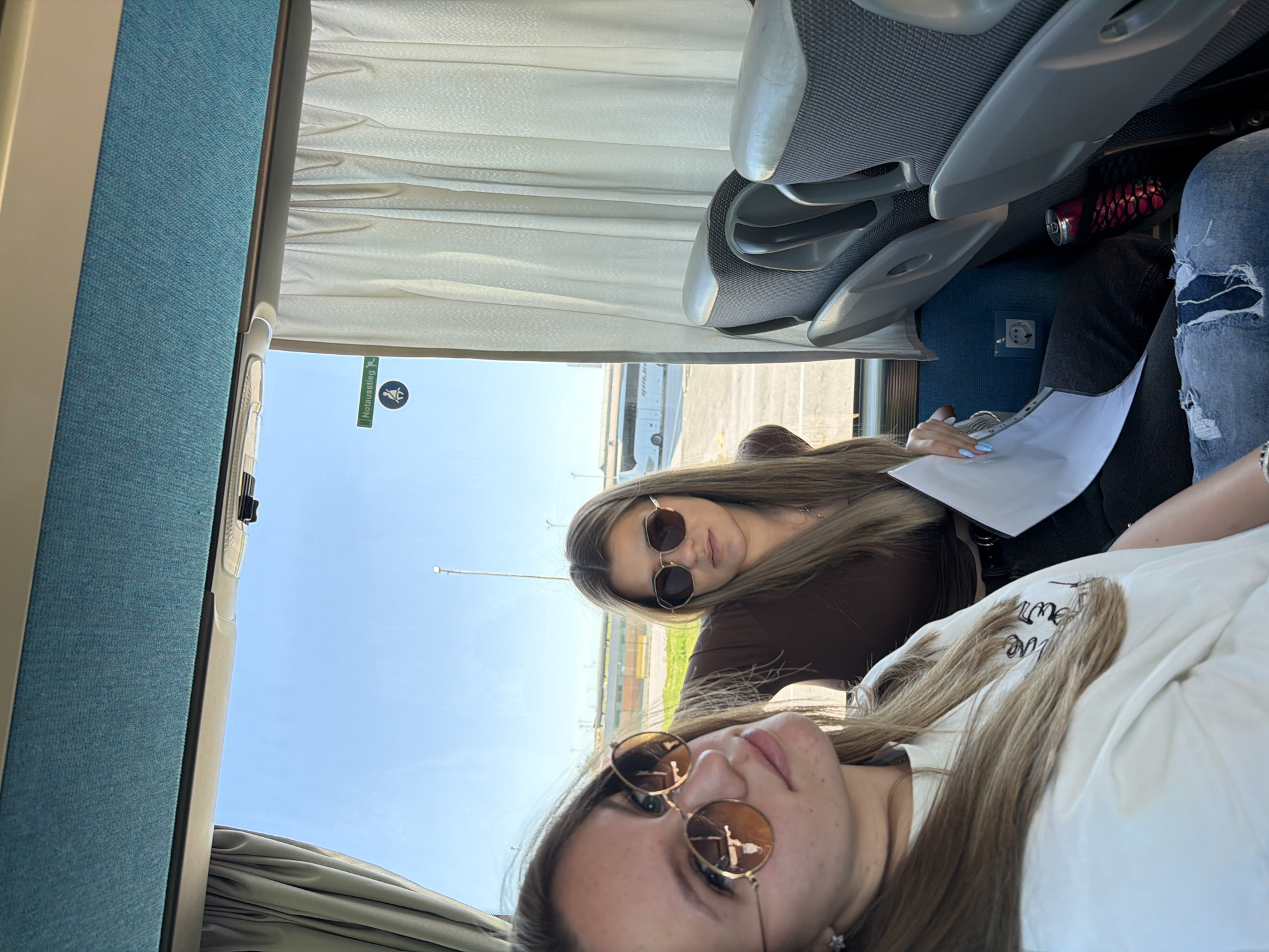 Two young women are sitting on a bus. The woman closer to the camera wears sunglasses and a white t-shirt. The other woman, sitting behind, also wears sunglasses and a brown shirt, and holds papers in her hand. The bus's window reveals a view of what appears to be an airport with planes. The location could be near Kapitan Andreevo, Bulgaria, based on the presence of a border crossing.