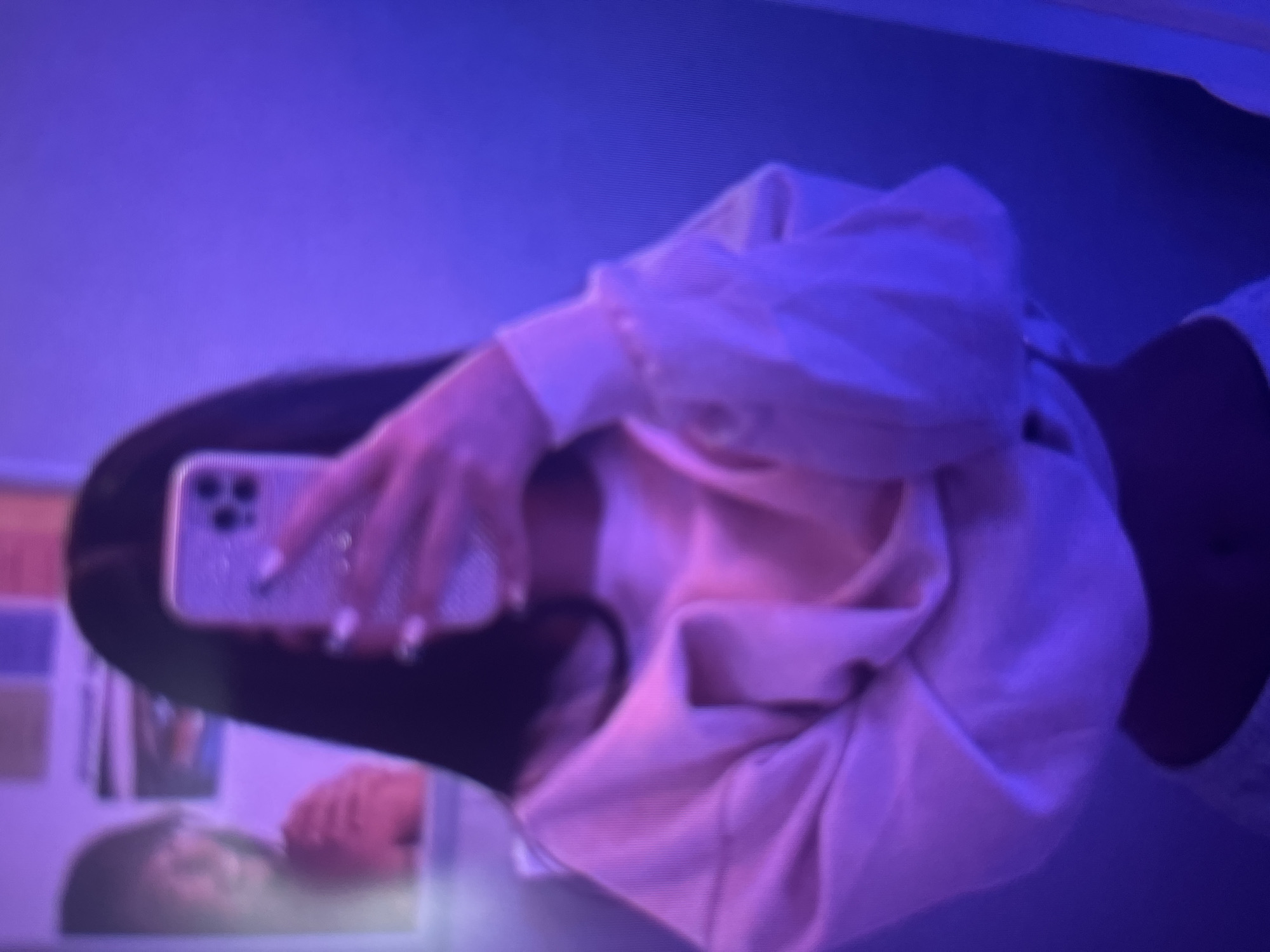 The image is a mirror selfie of a person, presented rotated 90 degrees clockwise. The individual, who has long dark hair, is holding a smartphone with a sparkling, possibly jeweled, case in their right hand, clearly visible with light-colored manicured nails. They are wearing a light-colored, likely beige or light pink, long-sleeved sweatshirt or hoodie. A sliver of their midriff and a white garment underneath the sweatshirt are visible.
The scene is set indoors, bathed in a strong purple and blue ambient light, suggesting specific mood lighting, such as from LED strips, or low light conditions typically associated with evening or nighttime. The reflection in the mirror, below the main subject's torso, includes a blurred image of a person's face, possibly the same individual, along with indistinct background elements that appear to be colorful items or objects. The image quality is somewhat grainy. There is no visible text. The person's emotions are not discernible.