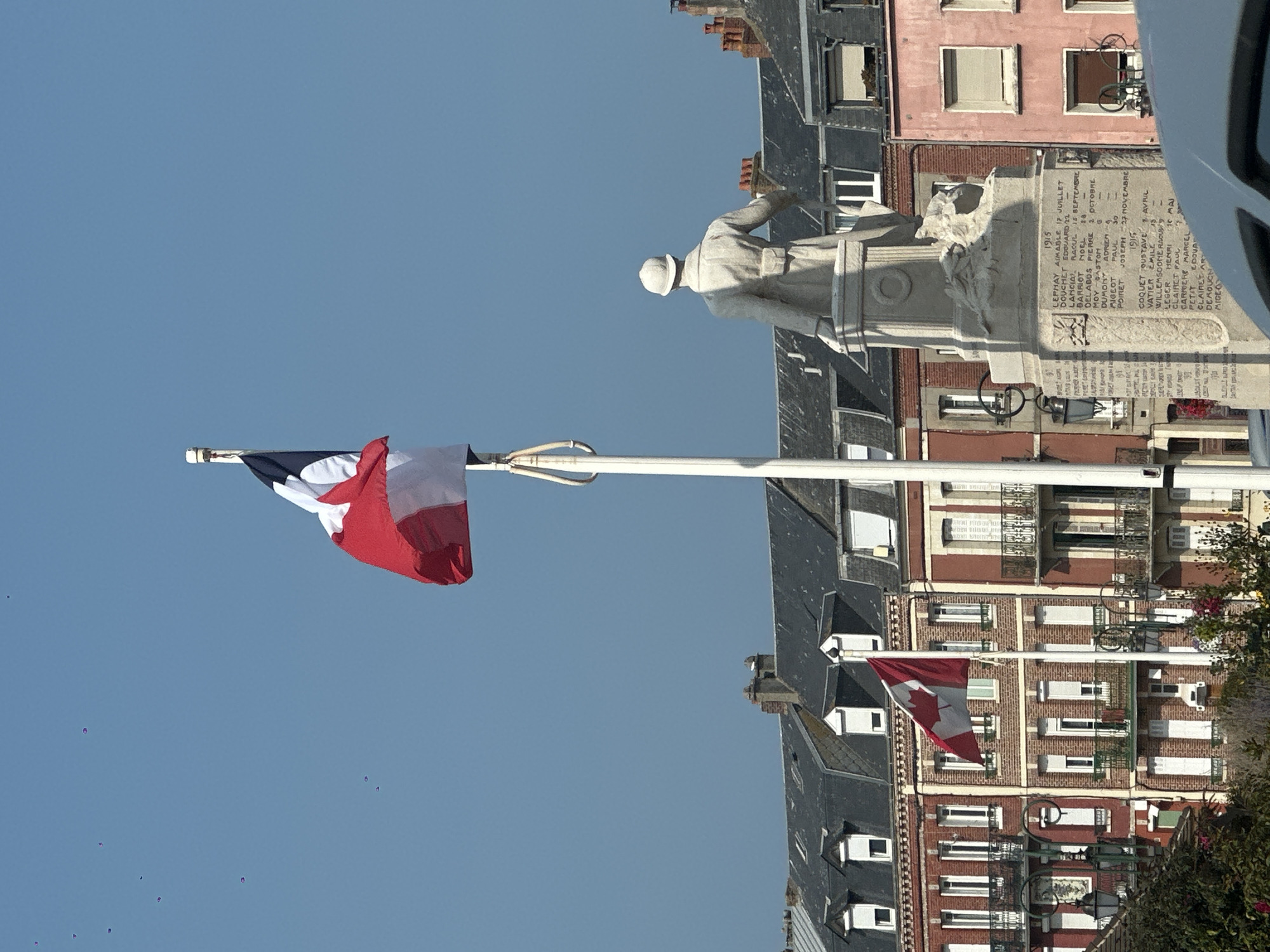 In Mers-les-Bains, France, a statue of a soldier stands in front of a row of multi-story buildings with red and pink facades. The soldier is depicted wearing a helmet and a trench coat, holding a rifle. A French flag flies on a tall flagpole to the left of the statue. In the background, a Canadian flag flies from another flagpole in front of a brick building. The sky is clear and blue, with a few small birds or objects flying in the distance. The scene appears to be a public square or memorial area, likely in a coastal town given the name Mers-les-Bains which translates to "Sea-Baths." The buildings show architectural details typical of European coastal towns, with pitched roofs, dormer windows, and wrought-iron balconies. The statue itself is a war memorial, with inscriptions carved into its base listing names and dates, suggesting a historical context related to military service. The overall atmosphere is one of remembrance and civic pride.