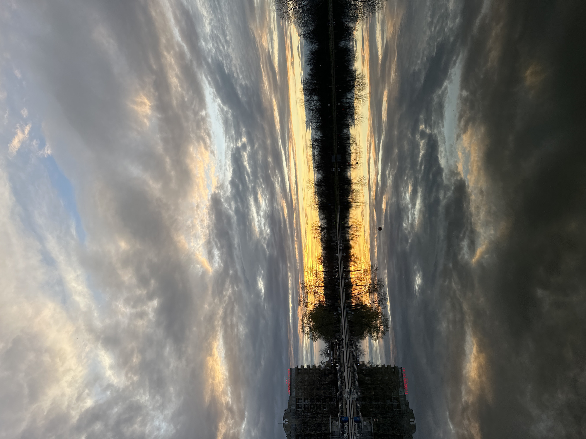 The image shows the reflection of the Plovdiv Rowing Canal in Bulgaria at sunset. The sky is filled with gray and white clouds tinged with gold from the setting sun. A line of trees borders the canal on both sides, reflected in the water. At the bottom of the image is the reflection of a building and a walkway. The image is rotated 180 degrees, making the building and walkway appear upside down.