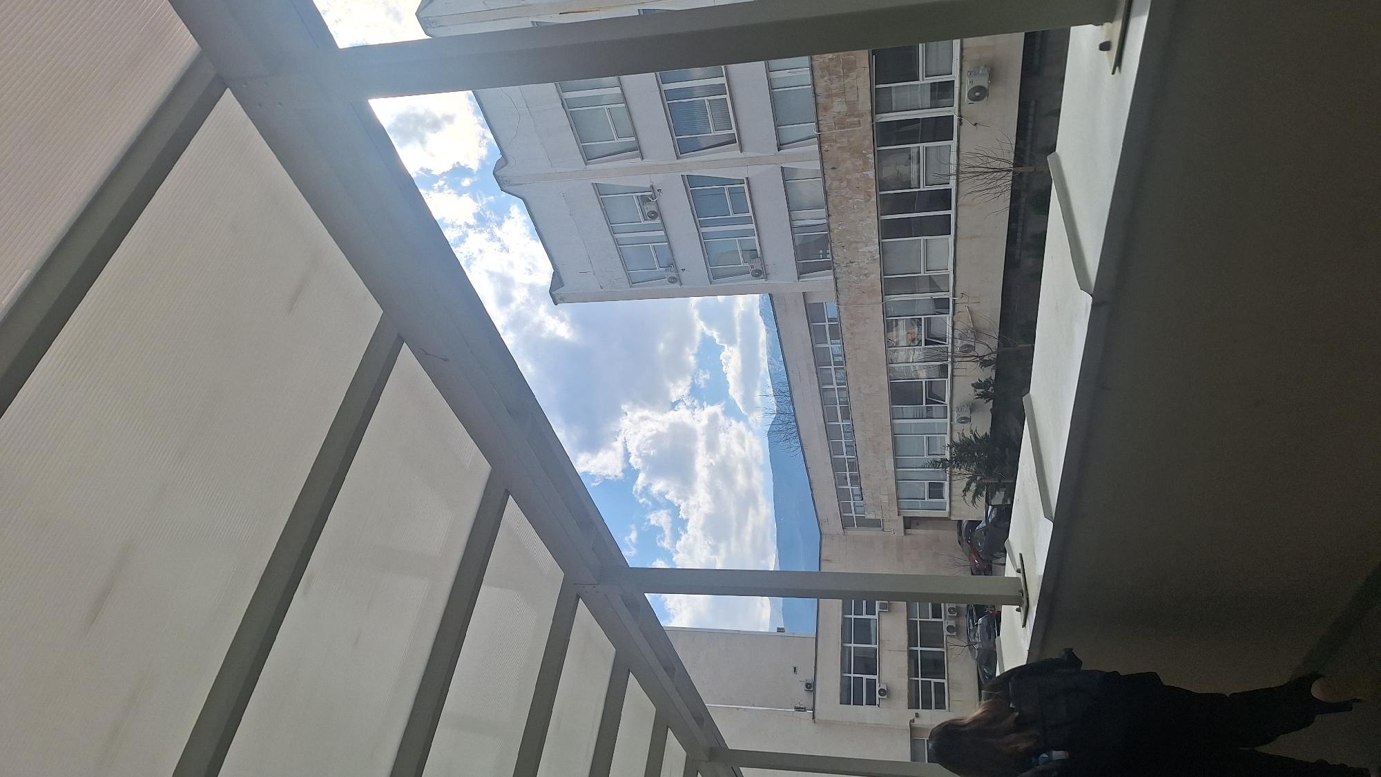 The image depicts an outdoor scene, likely from a walkway or balcony, framed by a translucent, corrugated polycarbonate canopy supported by grey metal beams. A single individual, seen from behind, is in the lower-left portion of the frame; this person has light brown hair and wears dark apparel, appearing to walk towards the left. A dark grey concrete parapet with a light-colored, multi-ribbed metal coping forms the immediate lower foreground.
Beyond the parapet, the campus of the Technical University of Sofia in Sofia, Bulgaria, is visible. The midground features multiple institutional buildings: to the left, a light-beige, multi-story building presents numerous rectangular windows and several wall-mounted air conditioning units. A taller, off-white, multi-story building with a distinctive stepped upper facade, similar windows, and additional air conditioning units is situated on the right. A lower, elongated structure with extensive fenestration connects these two primary buildings.
The background reveals a bright blue sky with scattered white cumulus clouds. The snow-capped Vitosha mountain range is prominently visible beyond the structures. Deciduous trees, currently bare, and some evergreen shrubs are sparsely distributed within the campus grounds. Several parked automobiles are partially discernible near the base of the left building. The overall scene suggests a university campus environment during a temperate or cold season.