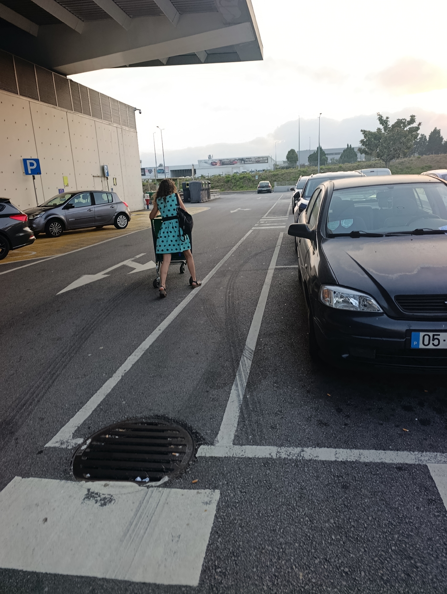 A woman in a polka dot dress pushes a shopping cart in a parking lot. Cars are parked in the background. The scene is outdoors, with a building on the left and a cloudy sky above. The image appears to be taken in Santa Maria da Feira, Portugal, based on the license plate of the car in the foreground which reads "05-".