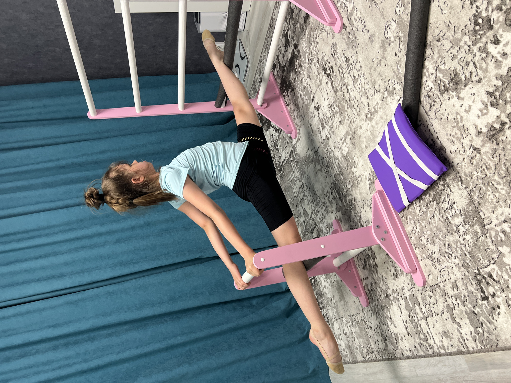 A young girl, around 10 years old, is captured performing a split on a pink and white gymnastics stretching tool in a room. She is wearing a light blue t-shirt and black shorts, with her hair tied back in a bun. Her left leg is extended upwards along a white ladder, while her right leg is supported by the stretching tool. She's leaning back, holding onto the apparatus with her hands. The room features a teal curtain in the background and a gray patterned carpet to the side. Next to the stretching tool on the carpet is a purple and white pillow. The image appears to be taken indoors, likely in a studio or home gym. While the exact location cannot be determined from the image, it may be somewhere in the city of Chaikovsky, Russia.