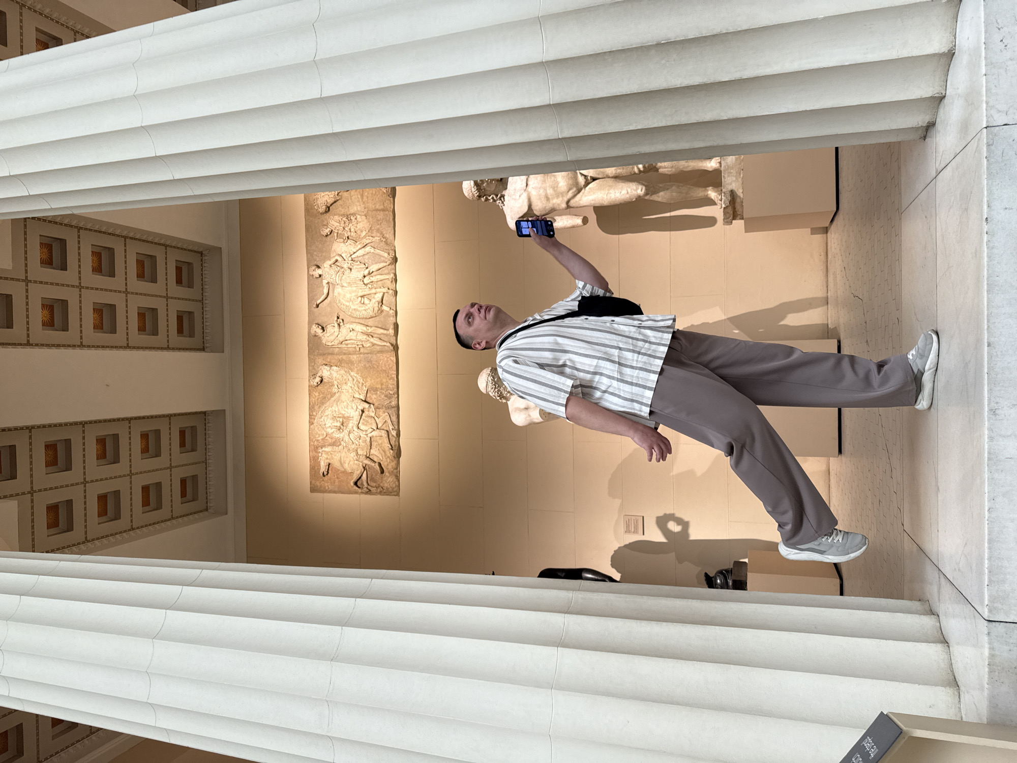 A man in a striped shirt and grey pants is photographing sculptures in a building with classical architecture. The building has fluted columns and the sculptures are mounted on walls, possibly friezes. The man is holding a smartphone up to take the picture. The location is not Moscow, Russia.