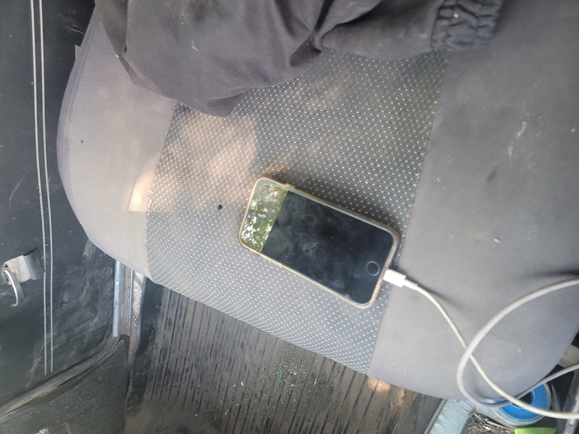 The image displays the interior of a vehicle, likely a car, in Pereshchepyne, Ukraine. A smartphone is positioned on the textured grey seat, connected to a white charging cable that trails across the seat and down towards the floor. The phone's screen is dark and shows reflections of greenery and what appears to be the car's interior. A dark fabric object, possibly clothing, is draped over the right side of the seat, partially obscuring the view of the seat's surface. The car's floor, visible to the left of the seat, is covered with black ribbed matting, showing evidence of dirt and debris. The car door panel is visible in the upper left corner, appearing to be made of dark plastic with metallic accents. The overall environment suggests a potentially untidy or well-used vehicle interior.