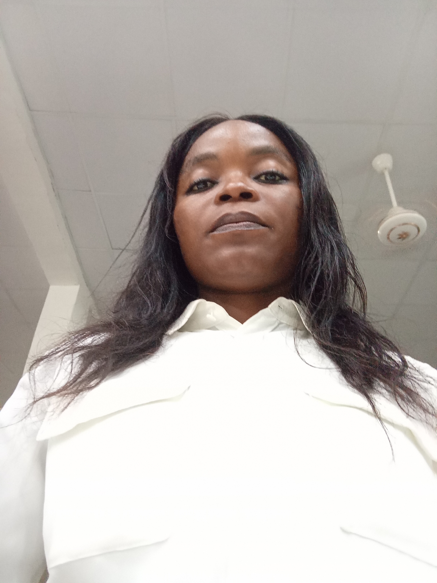 The image shows a low-angle, medium shot of a Black woman from the chest up. She has long, dark, wavy hair and is wearing a white collared shirt. Her eyes are open and looking directly at the camera. The background is an indoor ceiling with light fixtures and a fan. The scene appears to be a brightly lit room. No specific location in Kason, Nigeria can be determined from the visual information provided.
