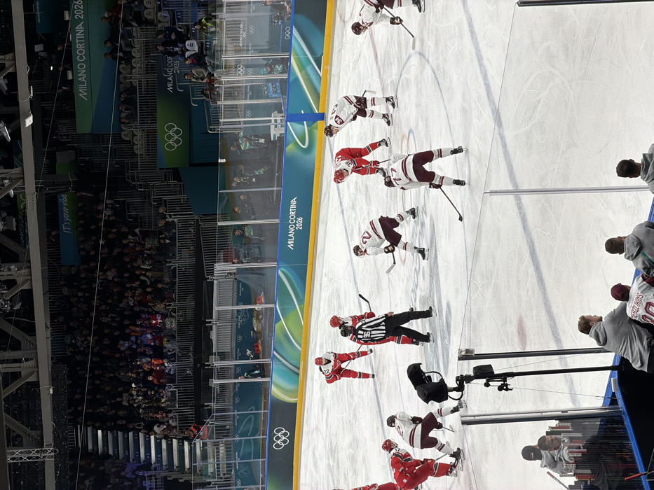 An ice hockey game is in progress at a venue advertising "Milano Cortina 2026" with the Olympic rings. Players in red and maroon uniforms face off, overseen by a referee. Spectators fill the stands, some partially obscured by the rink's plexiglass barrier. A banner reads "It's your vibe." Close to the barrier, one of the maroon players has the name "TERZLIKINS" and number "10" written on his back.