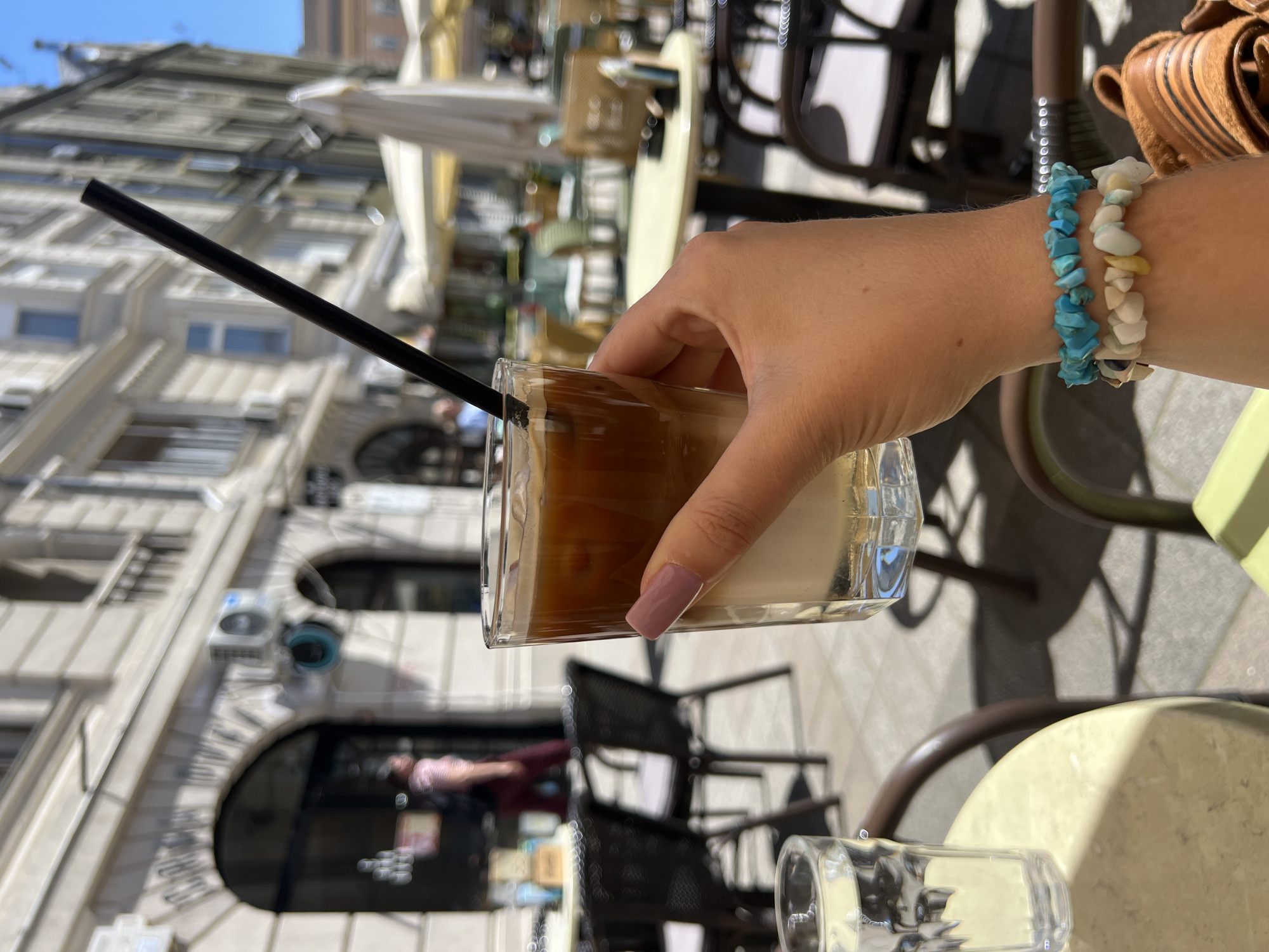 The image depicts a person holding a glass of layered coffee with a black straw while sitting at an outdoor cafe in Novi Sad, Serbia. The person is wearing two bracelets made of turquoise and white stones. The cafe setting includes wicker chairs, round tables, and other glassware on the table. In the background, the architecture of Novi Sad is visible, showing a building with arched windows and doors. The photo is taken on a sunny day.