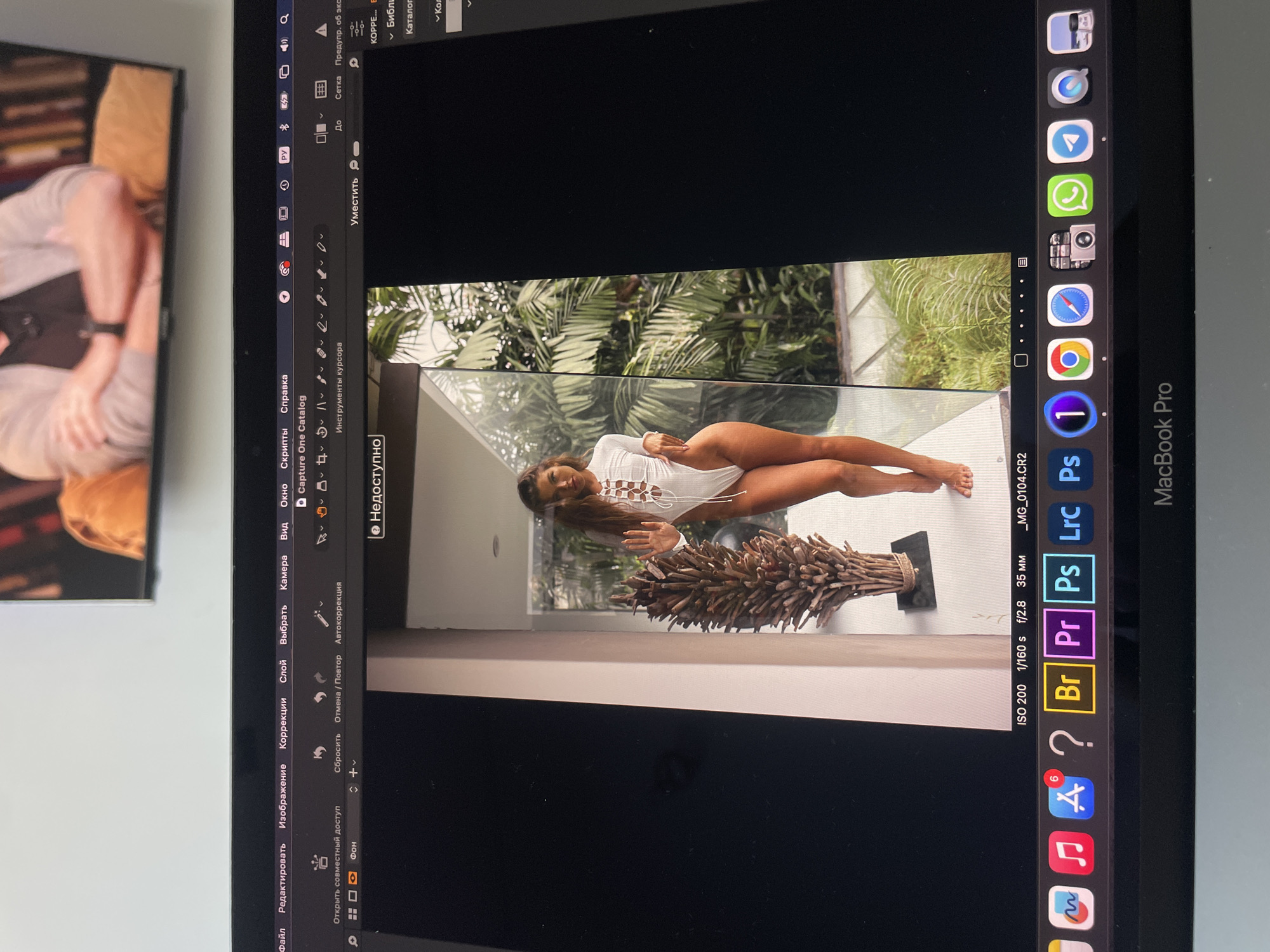 A MacBook Pro displays the Capture One photo editing program with an image of a woman in a white swimsuit lying horizontally on a ledge. The backdrop of the image is a lush green tropical setting. The woman is tanned and has long brown hair. In the foreground of the photo, a woven basket filled with sticks is visible. The MacBook Pro's screen also shows icons for programs in the dock, including Photoshop, Lightroom, and Bridge. The Capture One interface includes menus in Russian. To the left of the MacBook Pro's screen, the corner of a separate display shows a person seated, possibly watching television. The environment is indoors. The city of Kazan, Russia, is not directly visible in the image.