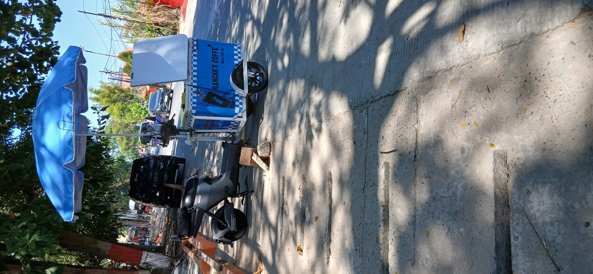An outdoor, daylight scene shows a mobile coffee cart parked on a concrete sidewalk next to a street. The cart is blue and white, with "RRANCAK'S COFFE" written on its side along with a stylized "R" logo. It has a white canopy and a white board at the top. A dark-colored electric scooter is parked to the left of the cart, propped up by several rocks. The street is visible in the background with several cars, including a black SUV, driving by. Trees with green foliage are overhead, casting dappled shadows on the ground. The environment appears to be an urban setting in Samarinda, Indonesia, based on visual cues such as the architecture and vehicles commonly seen in the region.