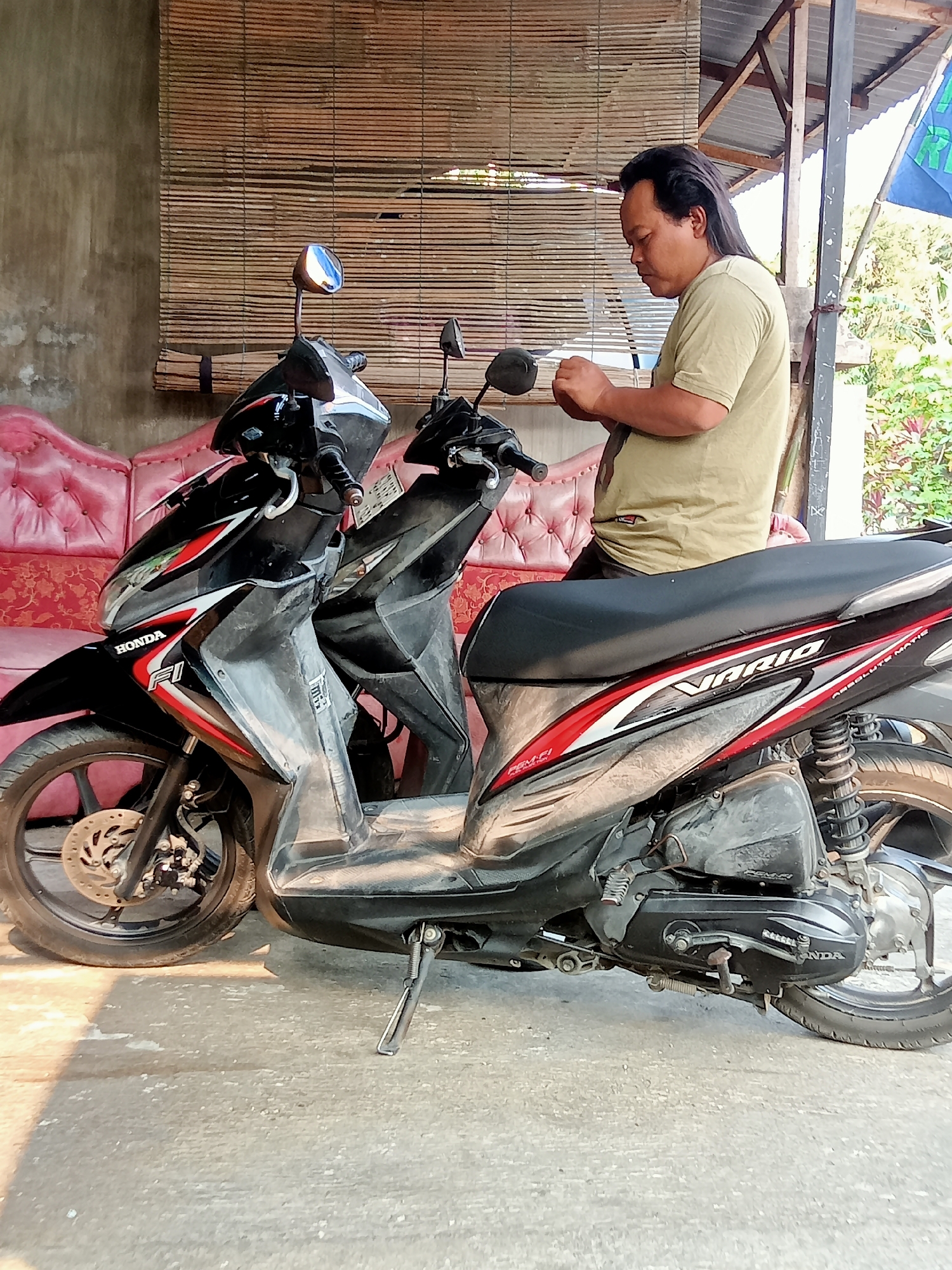 A medium-shot photograph captures a man in a light green t-shirt standing next to a black Honda Vario scooter in what appears to be an outdoor residential area. The man is facing away from the viewer, with his head turned to his left, and he is holding a small object in his hands, suggesting he is working on it or examining it. His long, dark hair is pulled back.
The scooter is parked on a concrete surface, with its kickstand down. It features red and white accents on its body. Behind the scooter, a pink tufted sofa is visible, and further back, a wall constructed of concrete and covered with a bamboo blind. The environment suggests a warm climate, with foliage visible in the background and sunlight casting shadows on the ground. The presence of a blue flag with white lettering can be seen to the right, slightly out of focus. The scene is likely in Majan, Indonesia, given the typical architectural style and the make of the scooter.