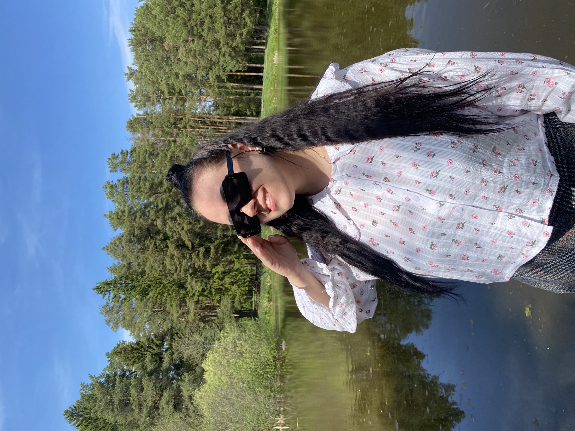 A woman with long, dark hair stands outdoors on what appears to be a dock or a bridge next to a body of water, possibly a lake or pond. She is wearing sunglasses, a light-colored blouse with a floral pattern, and a dark plaid skirt or pants. She is partially turned towards the camera, holding her sunglasses with her right hand and smiling.
The background includes a mix of evergreen and deciduous trees. The sky is blue, indicating it is daytime. The water reflects the trees and the sky.
Based on the information provided, this photo was taken in the city of Chaikovsky, Russia.