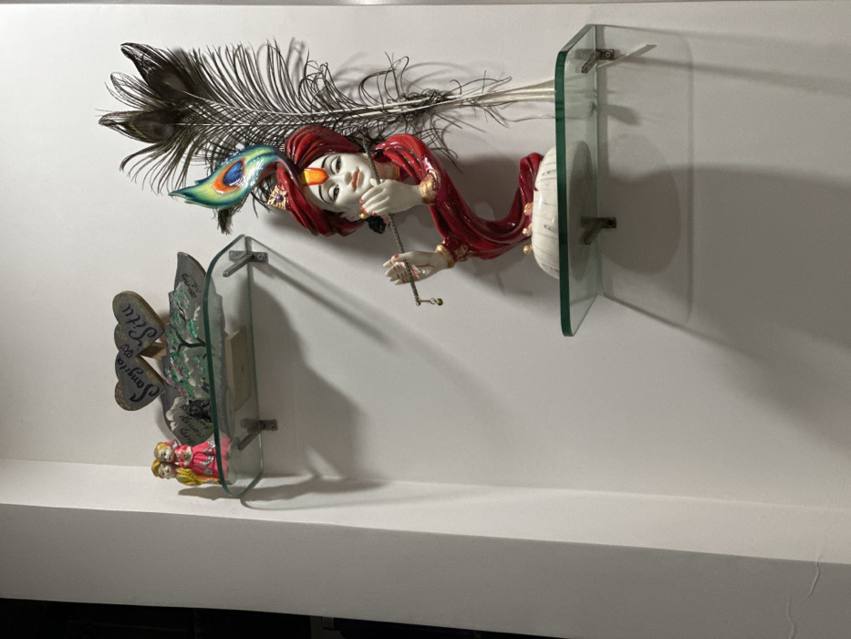 This image displays a section of a white interior wall in a residence or establishment located in Surat, India. Two clear glass shelves, supported by silver-colored metal brackets, are mounted on the wall.
The upper shelf holds a collection of decorative items: On the left, a pair of small, brightly colored figurines depict a male and female couple in traditional attire, with the female figure in a pink dress and the male in a yellow garment. Behind them, a green-tinted decorative piece, possibly resin, resembles a landscape with trees. To the right, two rustic, heart-shaped plaques are visible, one inscribed "SANGITA" and the other "SITU".
The lower shelf features a statue of Lord Krishna, rendered in white with prominent red and gold accents on his attire and turban. A vibrant, multi-colored peacock feather is integrated into his turban. The statue depicts Krishna playing a flute. To the right of the statue, a collection of natural, dark peacock feathers is arranged vertically against the wall. Shadows from the shelves and objects are cast onto the wall, suggesting illumination from an external or overhead light source.