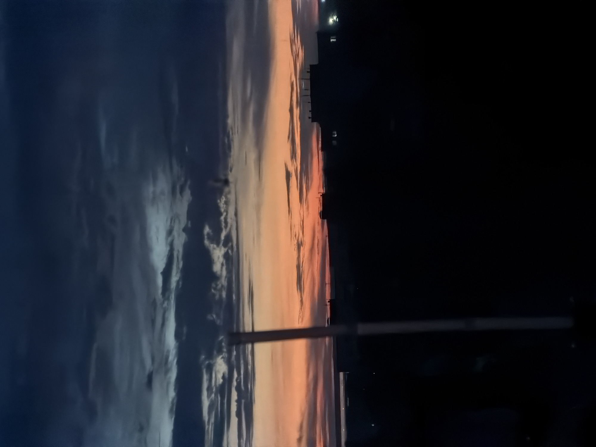 The image depicts a twilight scene in what is identified as Chaikovsky, Russia. The sky is divided between a dark blue, cloudy portion on the left and a gradient of orange and pink hues on the right, indicating either sunset or sunrise. Silhouetted buildings are visible in the far distance on the right side, close to the horizon. In the foreground, a vertical pole or beam is visible, interrupting the view of the sky in the center of the image. The area to the right of the pole is largely in shadow, with only faint details visible.