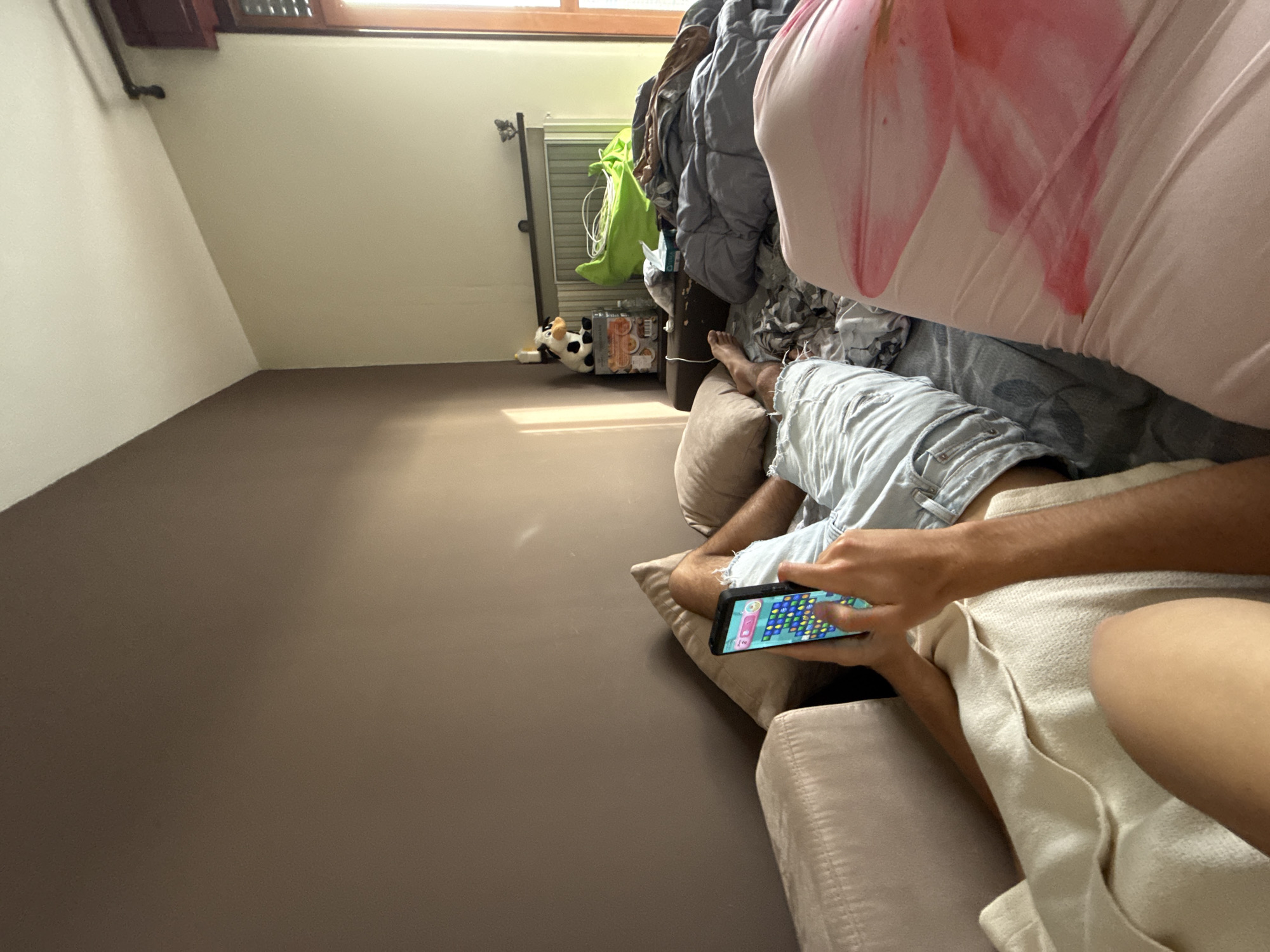 The image captures an indoor scene from a tilted perspective, showing a person relaxing on a sofa while engrossed in a smartphone game. The perspective makes the brown floor appear as a prominent wall in the lower left, meeting a lighter wall at an angle.
In the foreground, a person's arms and legs are visible as they sit or lie on a light-colored sofa. They wear light blue ripped denim shorts. Their hands are holding a black smartphone horizontally, actively playing a colorful match-3 puzzle game with visible scores or moves (e.g., "2", "4") on the screen. Another person's lower body, covered by a pink and white patterned fabric (possibly a blanket or part of clothing), is partially visible to the far right, suggesting another individual is also relaxing nearby.
The background of the room appears somewhat disheveled but cozy. A window along the top right allows natural light to stream in, illuminating parts of the room. Below the window, a pile of blankets and duvets, including gray and green fabrics, is stacked on what appears to be another sofa or bed. A small white and black spotted stuffed cow toy lies on the floor near some stacked boxes. The light from the window creates a bright strip on the floor/wall. The overall impression is one of casual relaxation in a home environment during the daytime.
