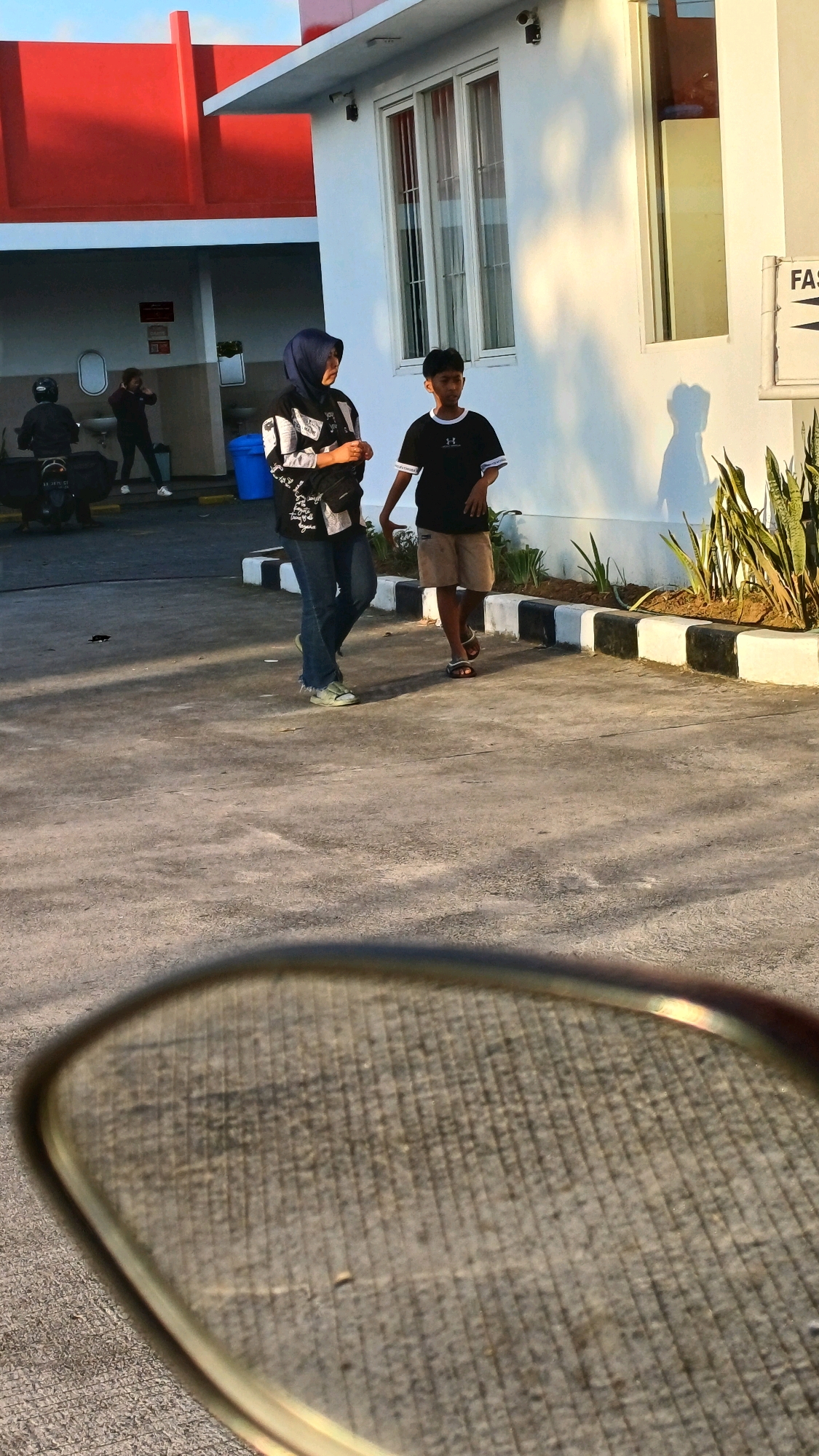 The image depicts an outdoor scene near a modern building with a white facade and a bright red overhang. Two individuals, a woman wearing a dark hijab and a patterned black and white long-sleeved shirt over blue jeans, and a young boy in a black t-shirt with a white logo and light brown shorts, are walking across a paved area. The woman is on the left, and the boy is to her right and slightly ahead. The woman is looking towards the boy, and the boy is looking forward. The paved area appears to be a walkway or parking lot, with a decorative border of black and white blocks and some greenery on the right side of the frame. In the background, a motorcycle with a rider is visible to the left, and further back, another person is standing near a sink area. A blue trash bin is also present. The light suggests it is either early morning or late afternoon, casting long shadows on the ground. The foreground is blurred, showing a reflective surface that could be a vehicle's side mirror. The overall environment suggests a public space, possibly near a shop or service area.