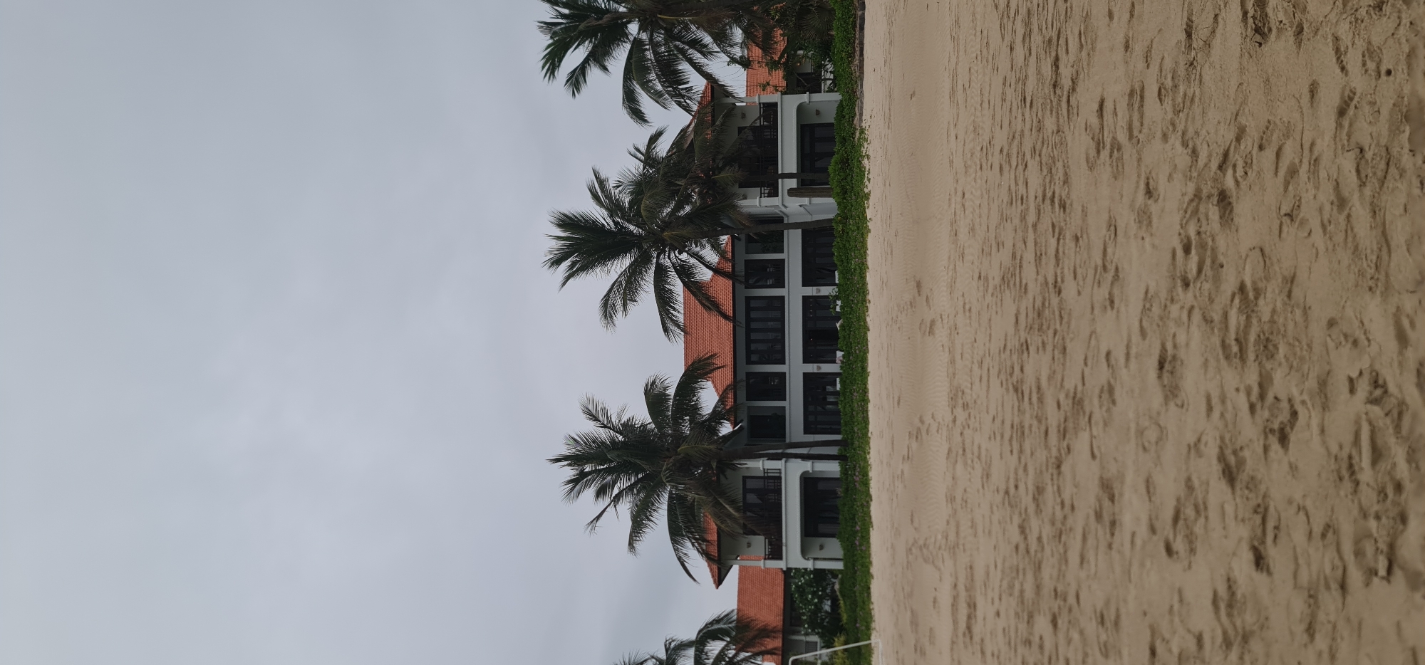 The image depicts a beach scene in Đà Nẵng, Vietnam. On the right side, there is light-colored sand with textured footprints. Adjacent to the sand is a thin strip of green vegetation. Beyond this, a two-story building with a red tile roof is visible. The building has white walls, dark window frames, and balconies. Palm trees are strategically placed in front of the building, partially obscuring the view. The sky is overcast and grey, indicating a cloudy day. No people are visible in the scene. The overall impression is of a tranquil beach setting with tropical vegetation and a coastal building.