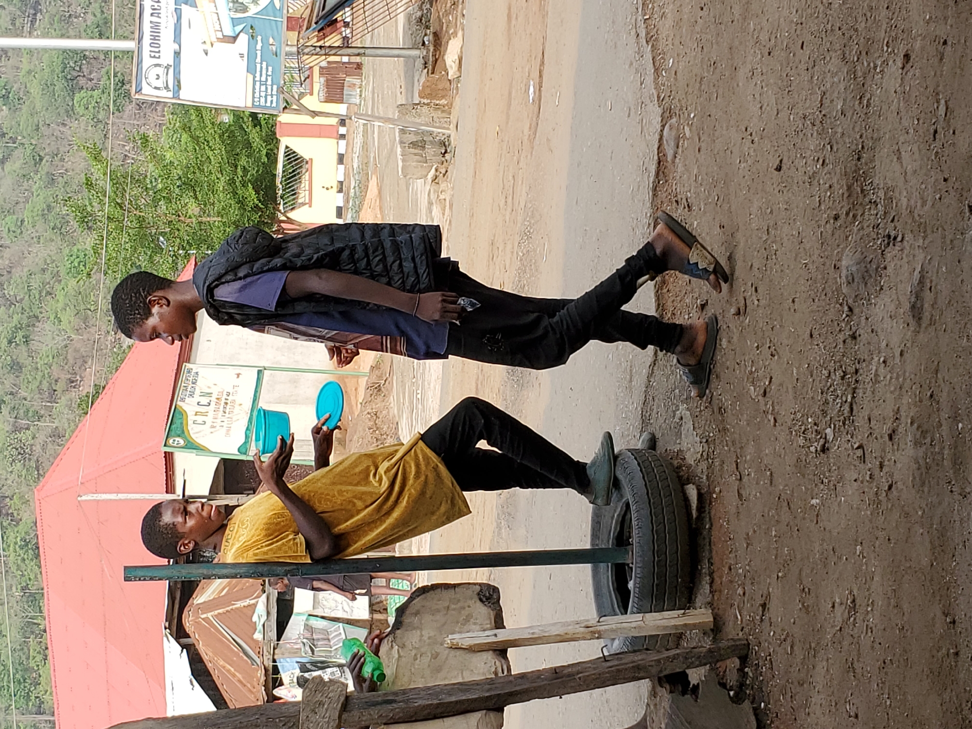 The image shows two young men on a street. One is seated on a wooden structure with a tire and metal bar, possibly a makeshift chair. He is wearing a yellow shirt and black pants, and he's holding a blue container. The other man stands behind him, wearing a dark vest over a blue shirt and black pants. In the background, there are buildings with signs reading "ELOHIM ACC" and "CRCN", indicating they are in a city with Christian presence. The OCR data mentions Mararaba, Nigeria, suggesting this is the location. The ground is a mix of dirt and paved road.