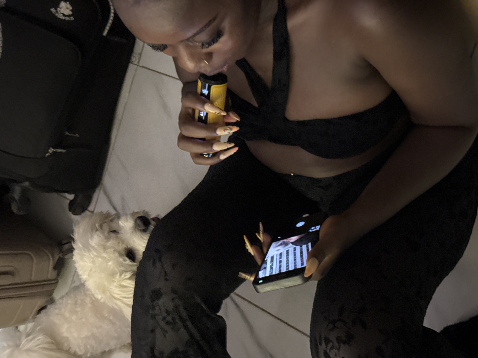 A medium shot captures a Black woman sitting on a tiled floor, with a small white dog to her left. She is holding a yellow vape device in her right hand to her mouth, and a smartphone in her left hand. She wears a black bra and black patterned leggings. Luggage is visible in the background. The scene appears to be indoors, possibly in a home or hotel room in Abuja, Nigeria.