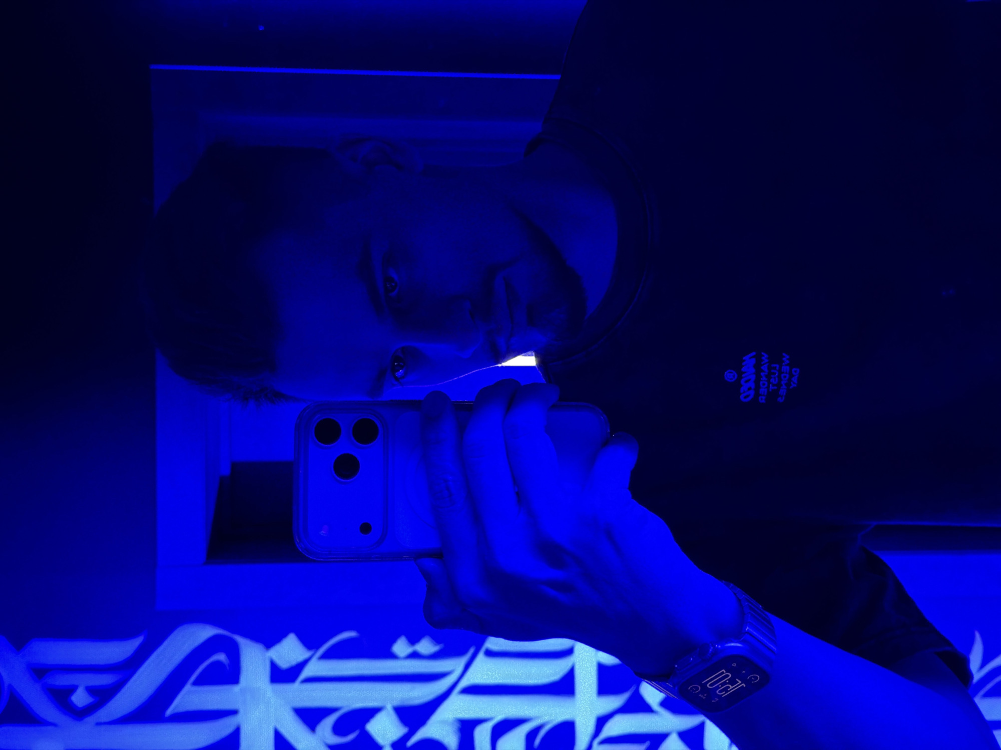 A male individual is taking a selfie in what appears to be a dimly lit or artificially lit environment with strong blue lighting. The man is holding an iPhone in his left hand, which is angled to capture his face and upper body. He is wearing a dark t-shirt with text and a logo on the chest. On his left wrist, he is wearing a watch with a visible digital display showing "12:01". The background includes white Arabic calligraphy. The location is not able to be determined from the image.