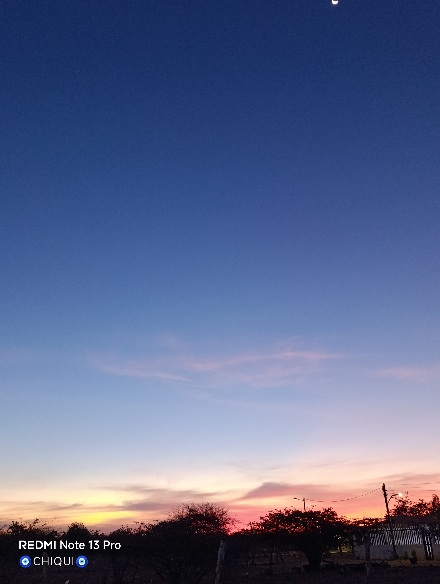 The image depicts a dusk or dawn landscape, captured in what appears to be a rural or semi-rural area. The sky is a gradient of deep blue at the top, transitioning to lighter blue, pink, and orange near the horizon. The moon is visible as a crescent in the upper right corner. The landscape includes trees, possibly shrubs, and fence posts silhouetted against the colorful sky. There are what seem to be buildings to the right. The image may have been taken in Cotoperíz, Venezuela based on the user's request. There is no indication of the presence of people in the scene.