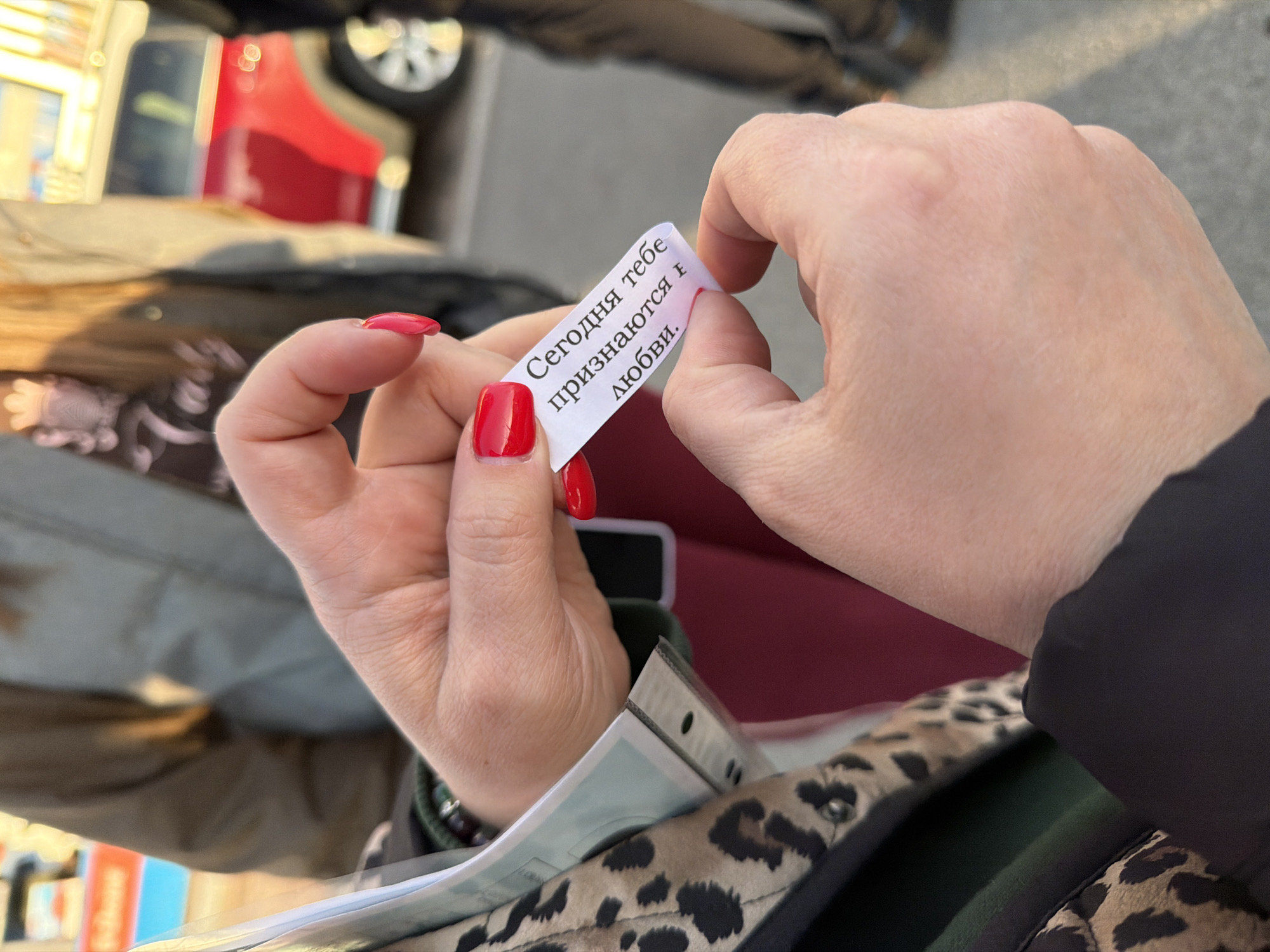 A close-up shot captures a person's hands holding a small, curved white slip of paper, commonly found in a fortune cookie. The left hand, adorned with bright red nail polish, grasps one end of the paper, while the right hand holds the other, presenting the message clearly. The text on the paper is in Cyrillic and reads: "Сегодня тебе признаются в любви." This translates to "Today someone will declare their love to you."
The background is softly blurred but suggests an outdoor, urban setting in Saint Petersburg, Russia. Partial views of a red car and other indistinct objects are visible, indicating a street or a public area. A section of a leopard-print bag or jacket is noticeable in the lower right corner, suggesting part of the person's attire. The scene is brightly lit by natural daylight, implying a clear and sunny day. The image focuses on the intimate moment of reading the fortune, conveying a sense of anticipation or personal reflection.
