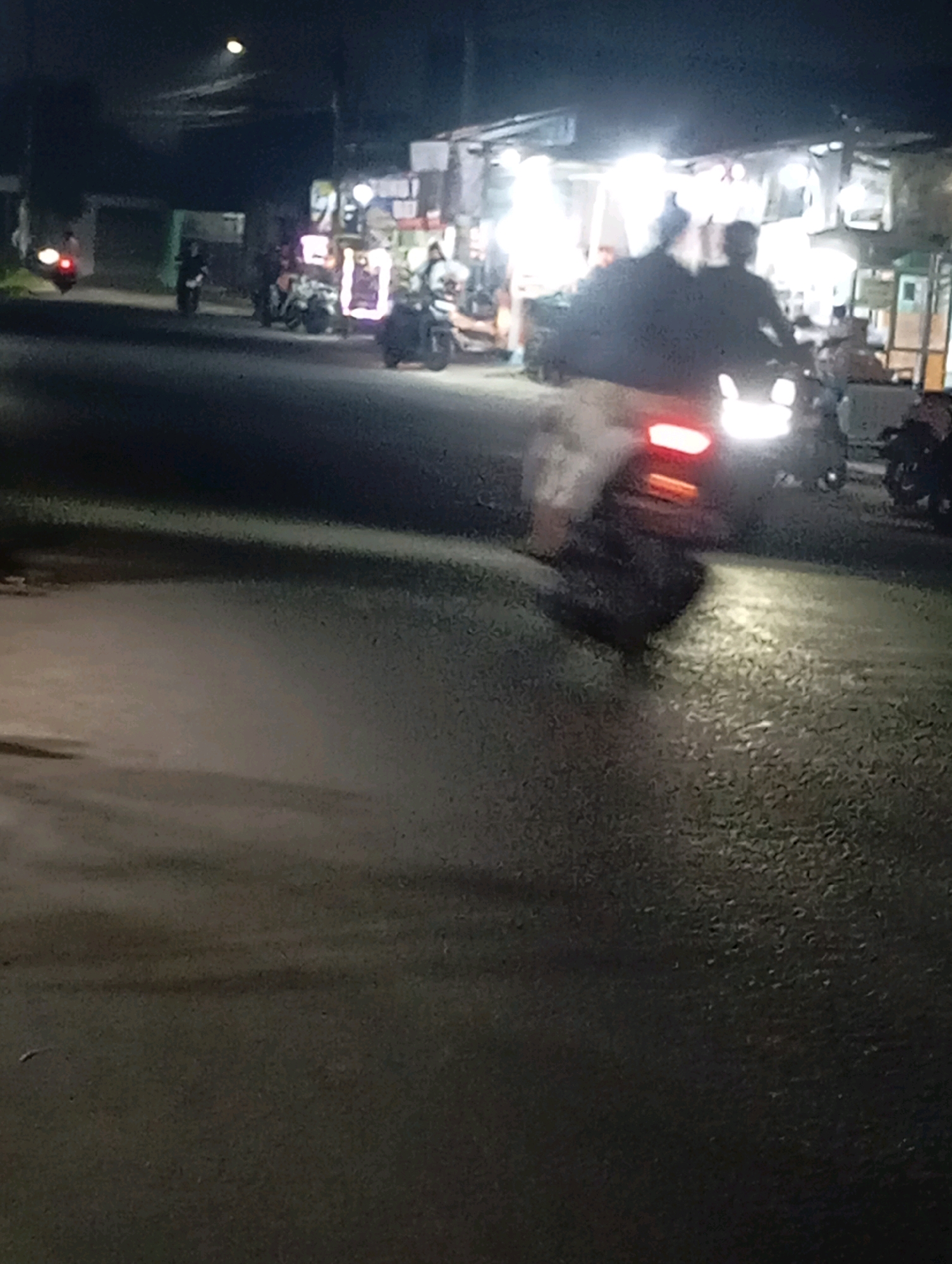 The image depicts a nighttime scene on a street in Babelan, Indonesia, likely a commercial area given the illuminated storefronts in the background. A motorcycle with two occupants is the primary focus, moving away from the viewer. The rider and passenger are silhouetted against the bright lights of the storefronts, making their features indistinguishable. The motorcycle's taillight is illuminated red, and its headlights cast a bright beam onto the asphalt road. The road surface appears wet or reflective, with shadows cast by the motorcycle and its occupants. In the background, several other motorcycles and figures are visible, some near illuminated stalls or shops. The overall atmosphere is dimly lit, with artificial lights from the businesses creating bright zones and the dark sky above.