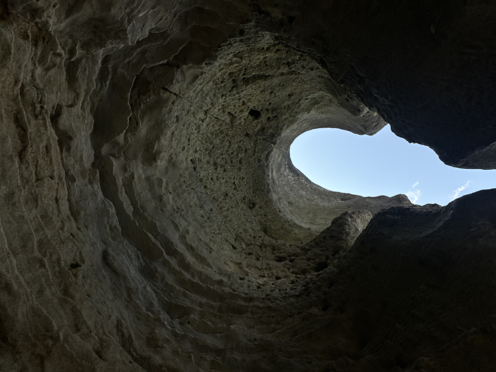 The image depicts the interior of a natural rock formation, possibly a cave or erosion-carved canyon, with a view looking upwards towards the sky. The rock walls are rough and textured, showing evidence of natural erosion with layered formations and numerous small holes or cavities. The sky visible through the opening is a clear blue with a few wispy clouds. The overall lighting suggests it is daytime, with the interior of the formation in shadow and the sky brightly lit. The scene is devoid of any people or man-made objects. This geological formation is characteristic of the landscapes found in the region of Aktau, Kazakhstan, known for its unique chalk and limestone cliffs and canyons.