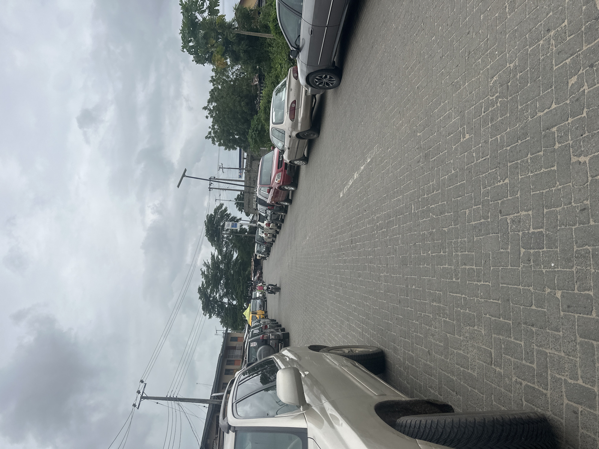 The image depicts a street scene in Badagry, Nigeria, under an overcast sky. The road is paved with interlocking grey bricks and is lined with parked cars on both sides. In the foreground, the rear portion of a silver SUV is visible on the left. Further down the street, on the right, a row of cars including a grey sedan, a tan sedan, and a red SUV are parked. On the left side of the road, a line of vehicles includes a black SUV and a yellow van. In the middle of the road, a person on a motorcycle is riding away from the camera, wearing a red shirt and a helmet. The background features buildings, trees, and utility poles with overhead wires. The overall impression is a typical urban street with moderate traffic and parked vehicles.