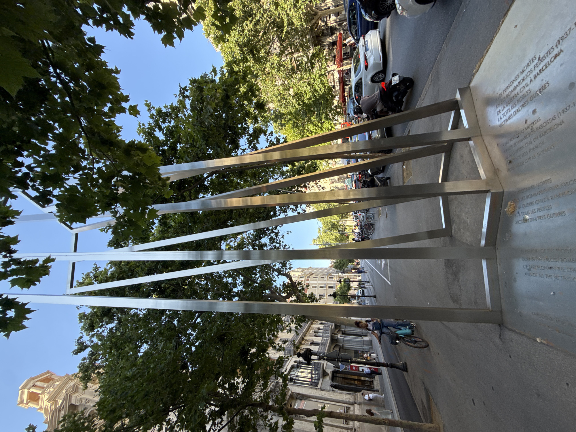 The image captures a street scene in Barcelona, Spain, featuring a metallic monument composed of vertically arranged bars. The monument bears inscribed text in Catalan, Spanish, and English, referring to the victims of fascist bombings during the Spanish Civil War (1937-1939) in Barcelona. The monument is positioned on a street lined with buildings and trees, with pedestrians and cyclists in view. Parked vehicles and motorcycles are visible along the curb. The sky is clear and blue. The photograph is taken from a low angle, emphasizing the height of the monument and incorporating the surrounding trees into the composition.