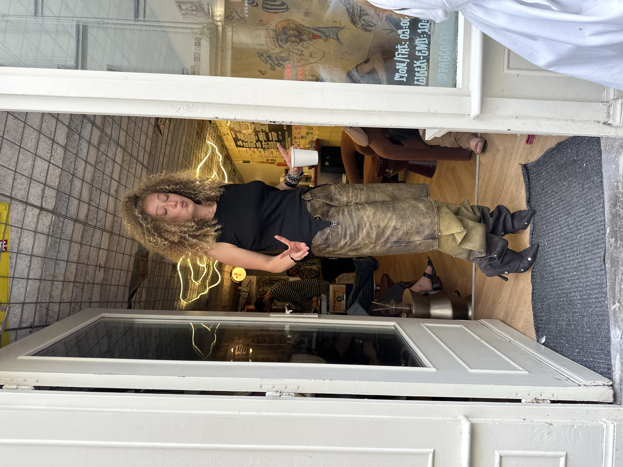 A woman with curly hair, a black shirt, leather-like pants, and boots is standing in the doorway of a business. She is holding a white cup and gesturing with her hand. Inside the business, there are other people, including a person in a striped shirt. Neon lights decorate the wall. A sign on the window lists the business hours and the social media handle "@PAGCOFFEE." The business appears to be in Paris, France, judging from the style of the building and street. The doorway is framed by white-painted wood.