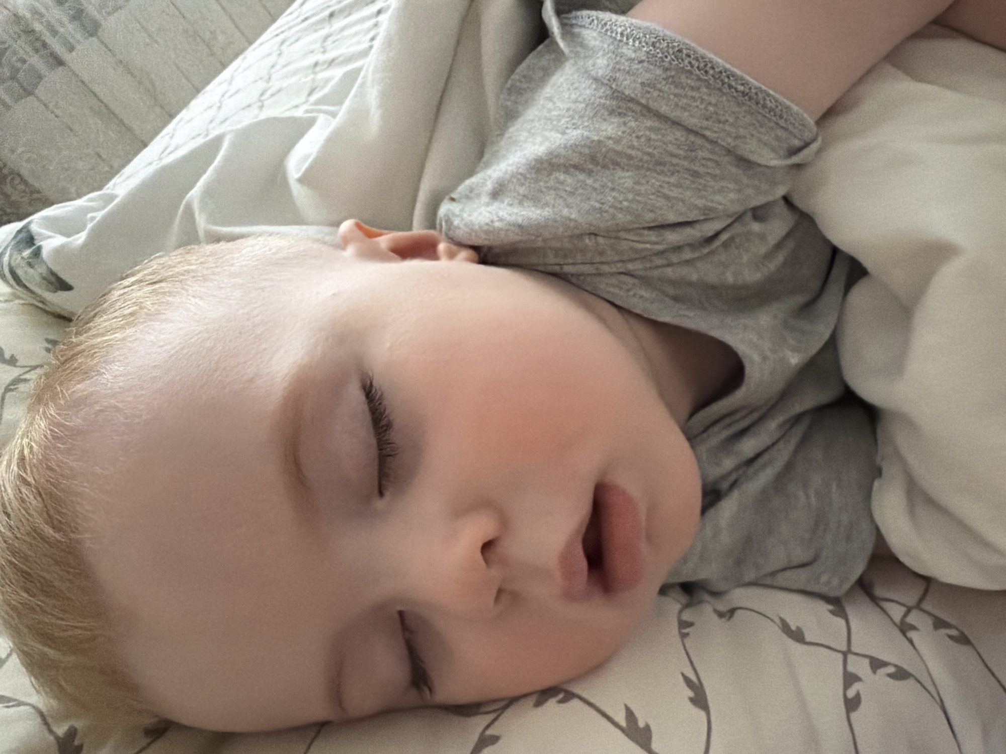 A close-up shot shows a sleeping infant with fair skin and light blonde hair. The child is lying on a patterned bedsheet with gray and white tones. The infant is partially covered by a gray t-shirt and a white blanket. The child's face is turned slightly to the side, with eyes closed and mouth slightly open. No information indicates the photograph was taken in Votkinsk, Russia.