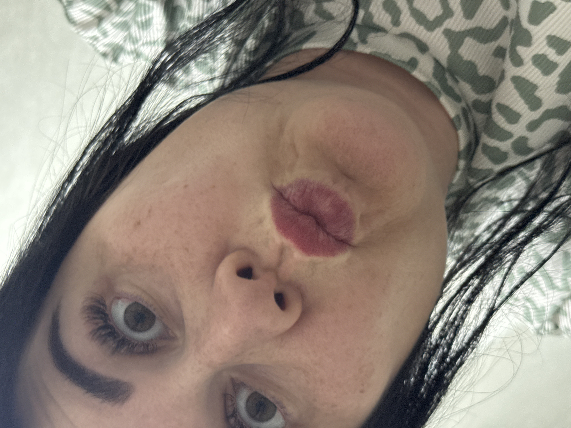 A selfie of a woman with fair skin, dark eyebrows, and dark hair, is shot from a low angle. She is making a duck face with lipstick on. The background includes a white surface and a patterned fabric in light green and white. The location of the image is unknown and there is no evidence to indicate this image was taken in Zav'yalovo, Russia.