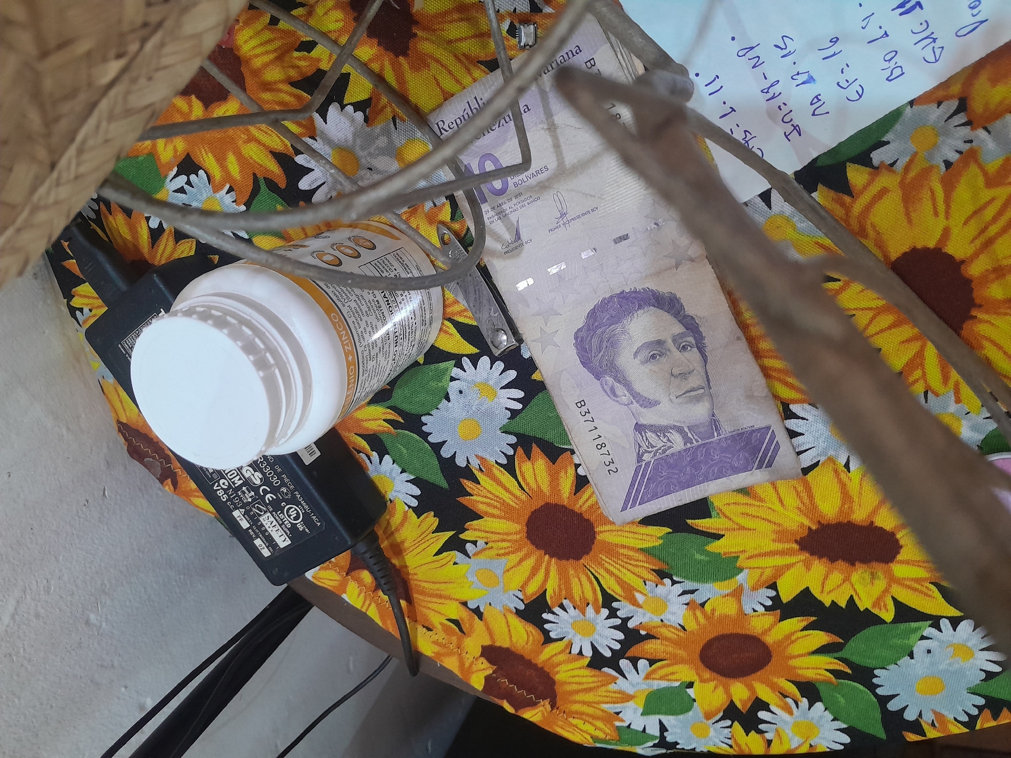 The image depicts a close-up overhead view of a table covered with a black fabric featuring a repeating pattern of sunflowers and daisies. On the table are several objects: a white plastic bottle with a white cap, a black power adapter with various certifications and markings, and a Venezuelan banknote. The banknote, a 10 Bolivar note, is predominantly purple and features a portrait of Simón Bolívar. Inscribed on the banknote are "República Bolivariana de Venezuela" and a date "29 DE ABRIL DE 2013".
Also visible is a small, metallic object, possibly a nail clipper or similar tool, placed near the bottle. To the right of the banknote, a white piece of paper with handwritten blue text is partially visible. The handwriting appears to be a list of alphanumeric characters and numbers, such as "IN=19-NP", "EF=16", and "Blo 1.4".
The background consists of a white wall and a portion of a wicker-like basket. The lighting suggests an indoor environment. The image does not provide any specific details that would allow for the determination of its location within the city of Santa Ana, Venezuela.